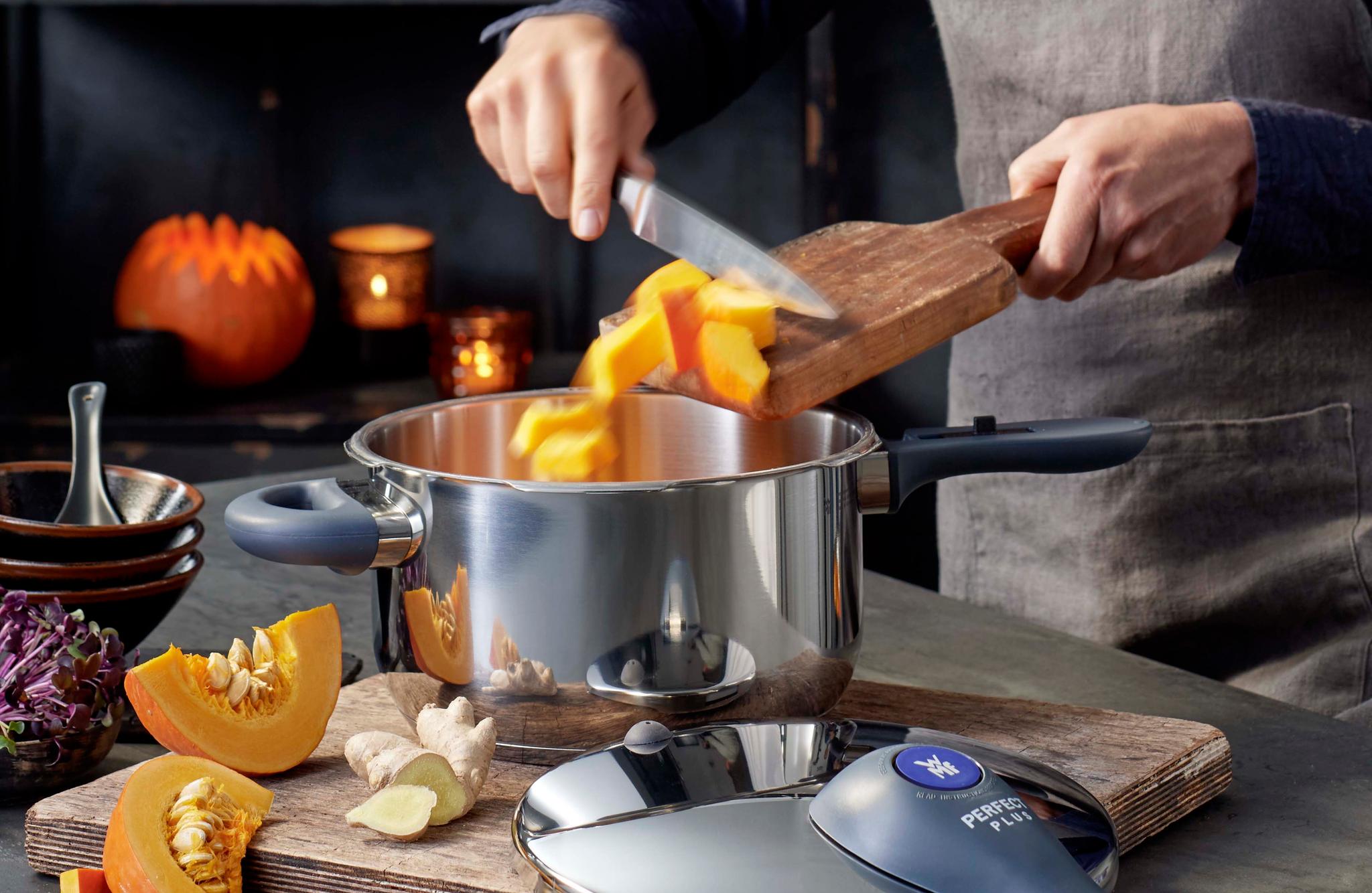 Lähikuva kokin käsivarresta puisella keittiön tasolla vasaroitujen ruostumattomasta teräksestä valmistettujen keittoastioiden vieressä.