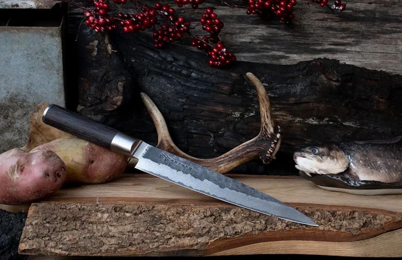 Sujihiki en lång kökskniv från Japan på en skärbräda med rotfrukter och lingon