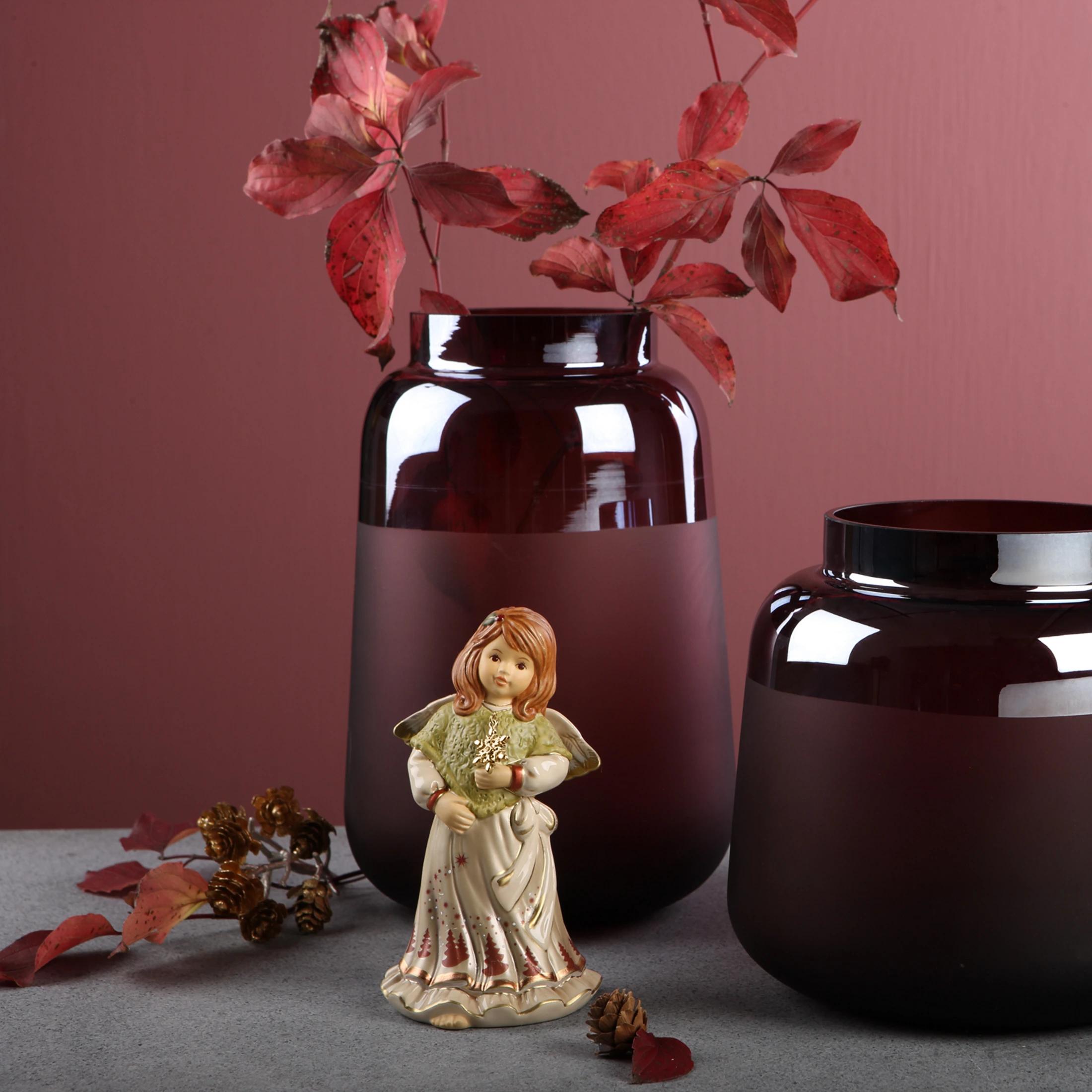 A ceramic angel figurine holding a snowflake, positioned in front of two frosted burgundy vases, with scattered autumn leaves and pinecones.