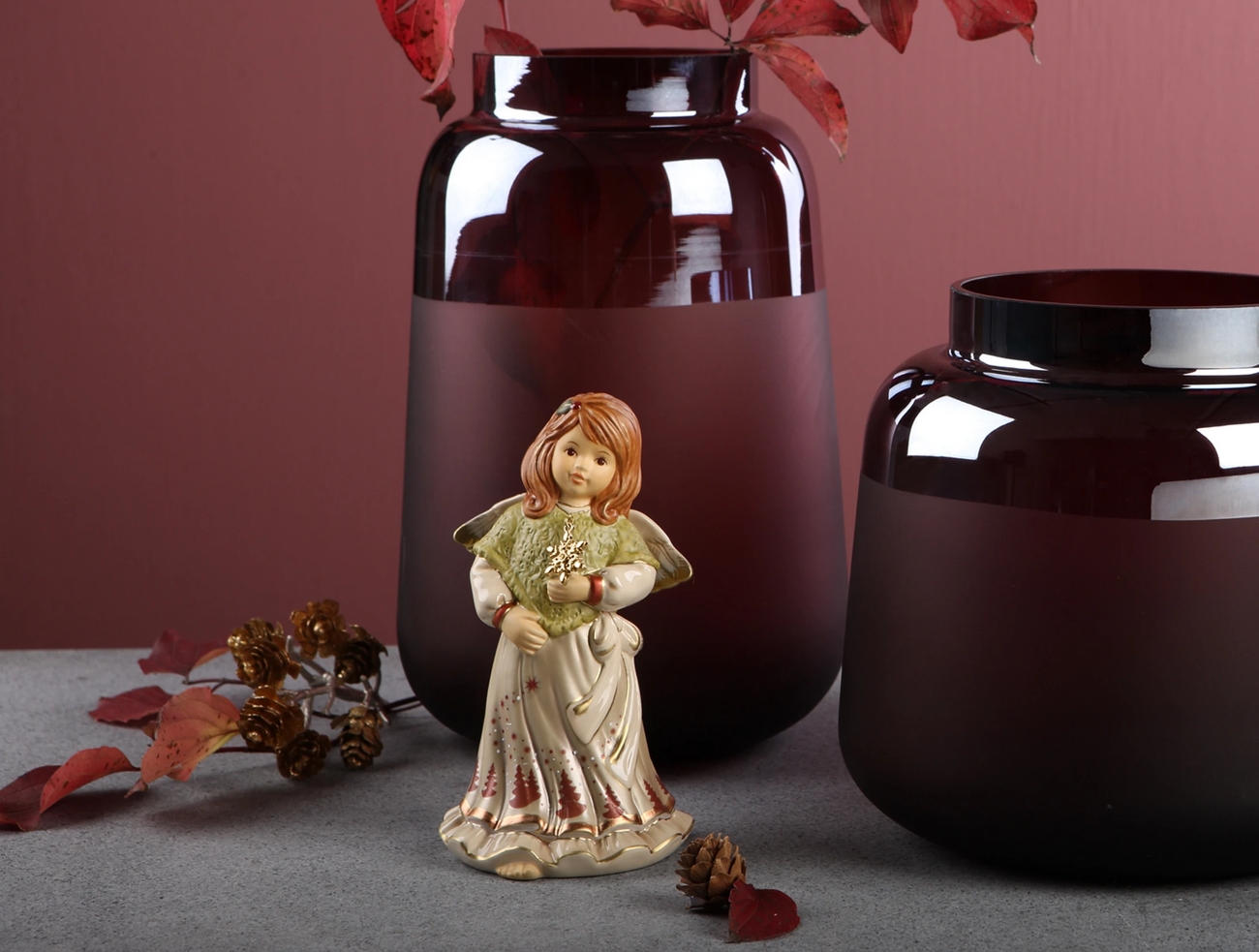 A ceramic angel figurine holding a snowflake, positioned in front of two frosted burgundy vases, with scattered autumn leaves and pinecones.