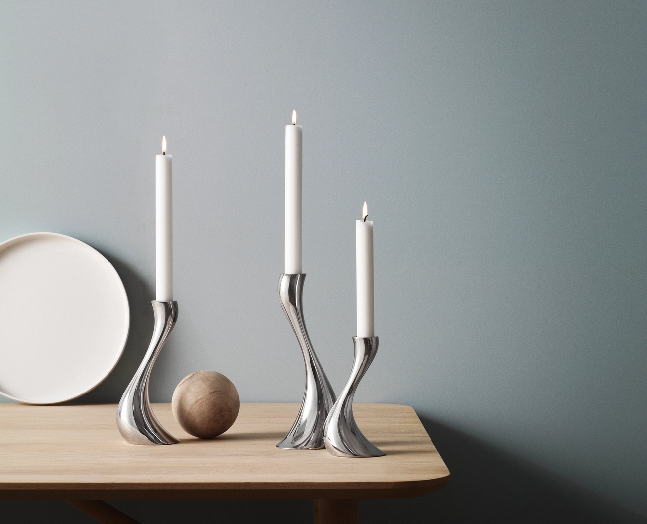 Tre silverfärgade, böjda ljusstakar med tända vita ljus på ett ljust träbord mot en blå vägg.