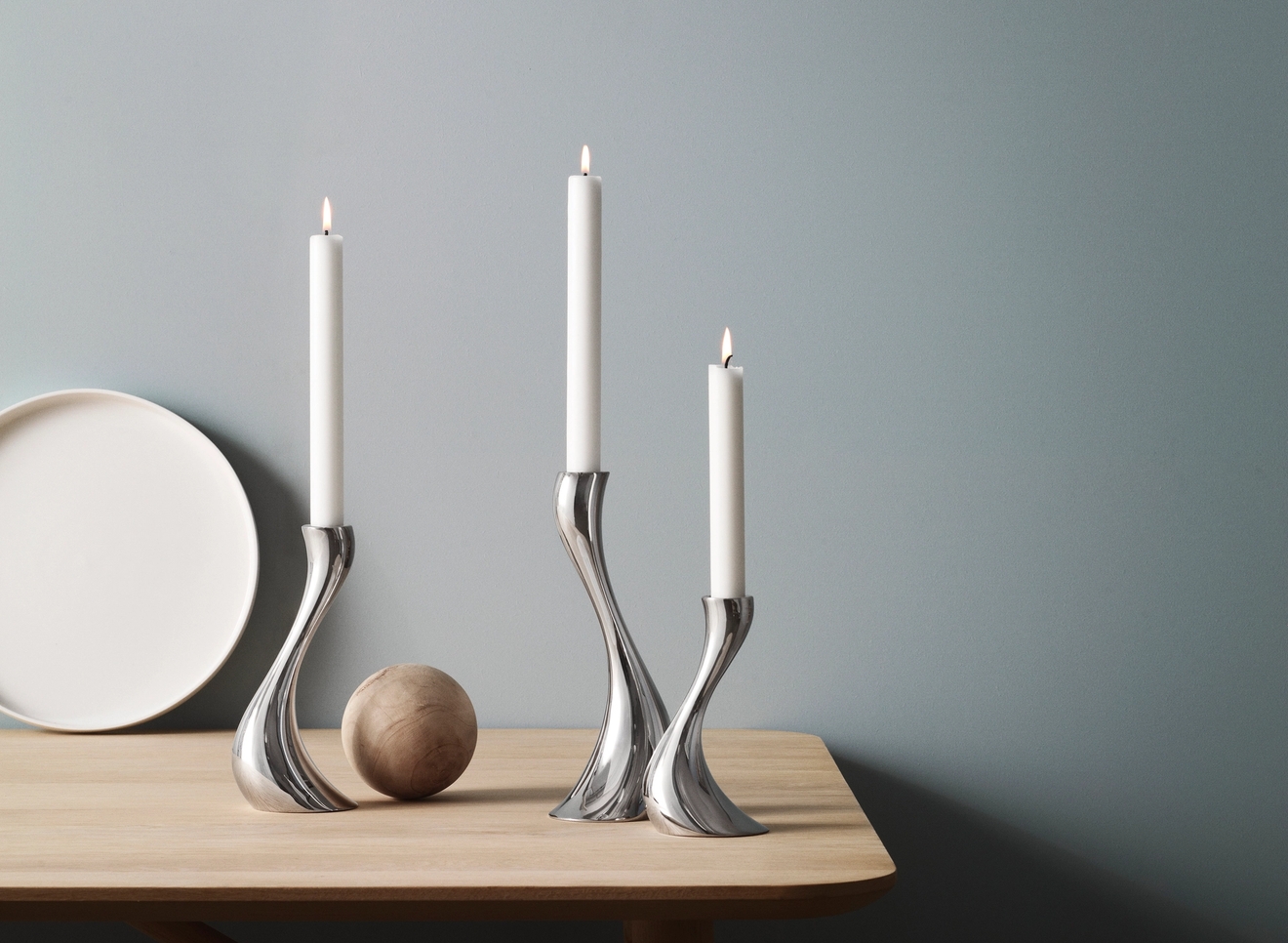 Tre silverfärgade, böjda ljusstakar med tända vita ljus på ett ljust träbord mot en blå vägg.