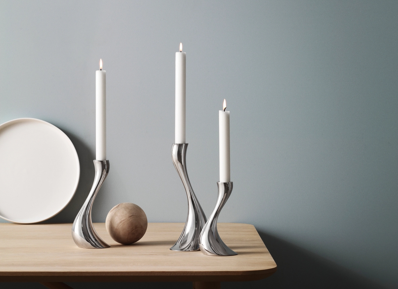 Tre sølvfargede, buede lysestaker med tente hvite lys på et lyst trebord mot en blå vegg.