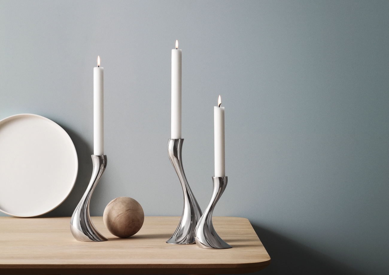 Tre sølvfargede, buede lysestaker med tente hvite lys på et lyst trebord mot en blå vegg.