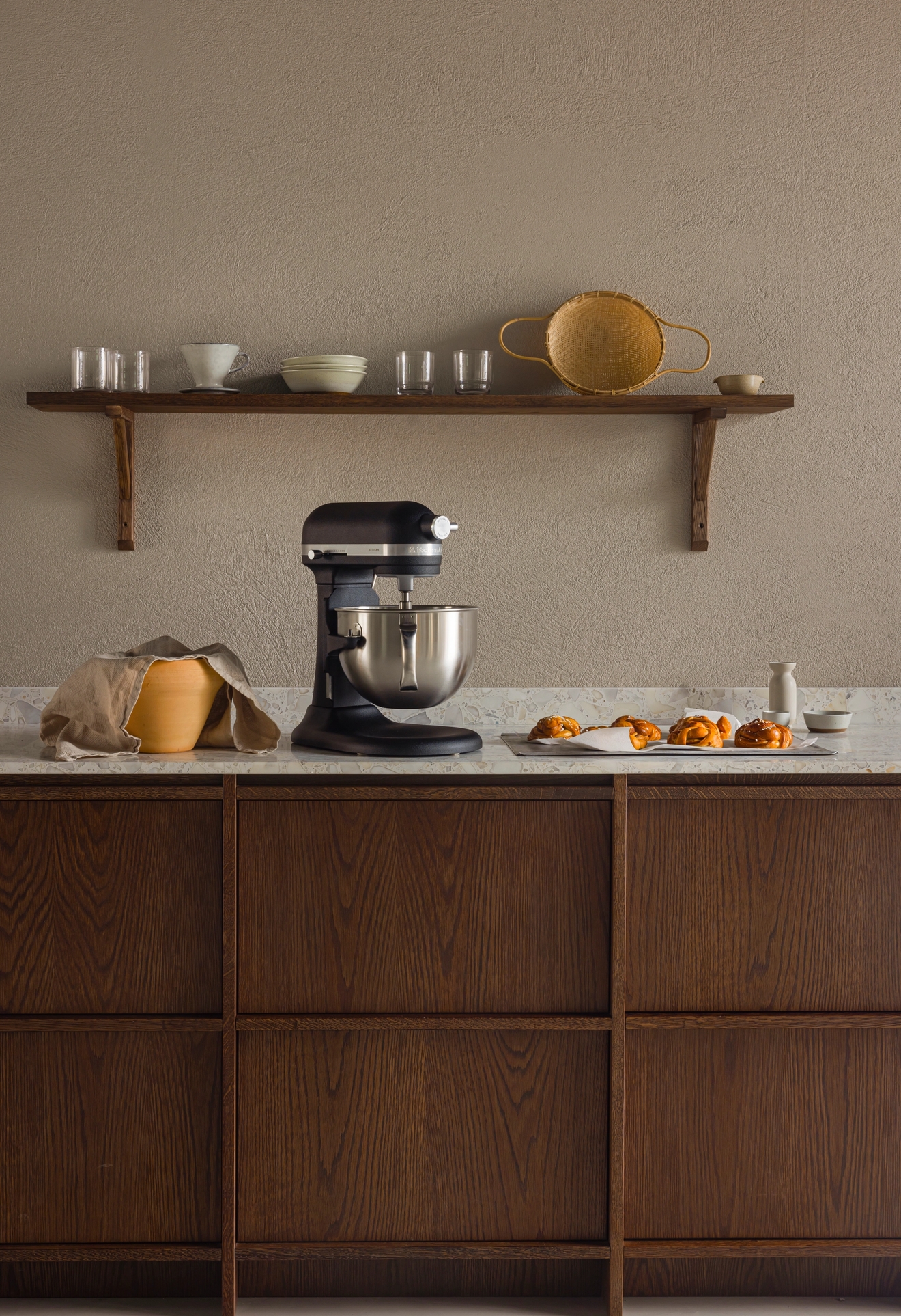 Keittiö, jossa on tummat puukaapit, vaalea työtaso, musta yleiskone, oranssi kulho ja tarjotin leivonnaisia. Yllä neutraalilla seinällä roikkuu puinen hylly, jossa on astioita