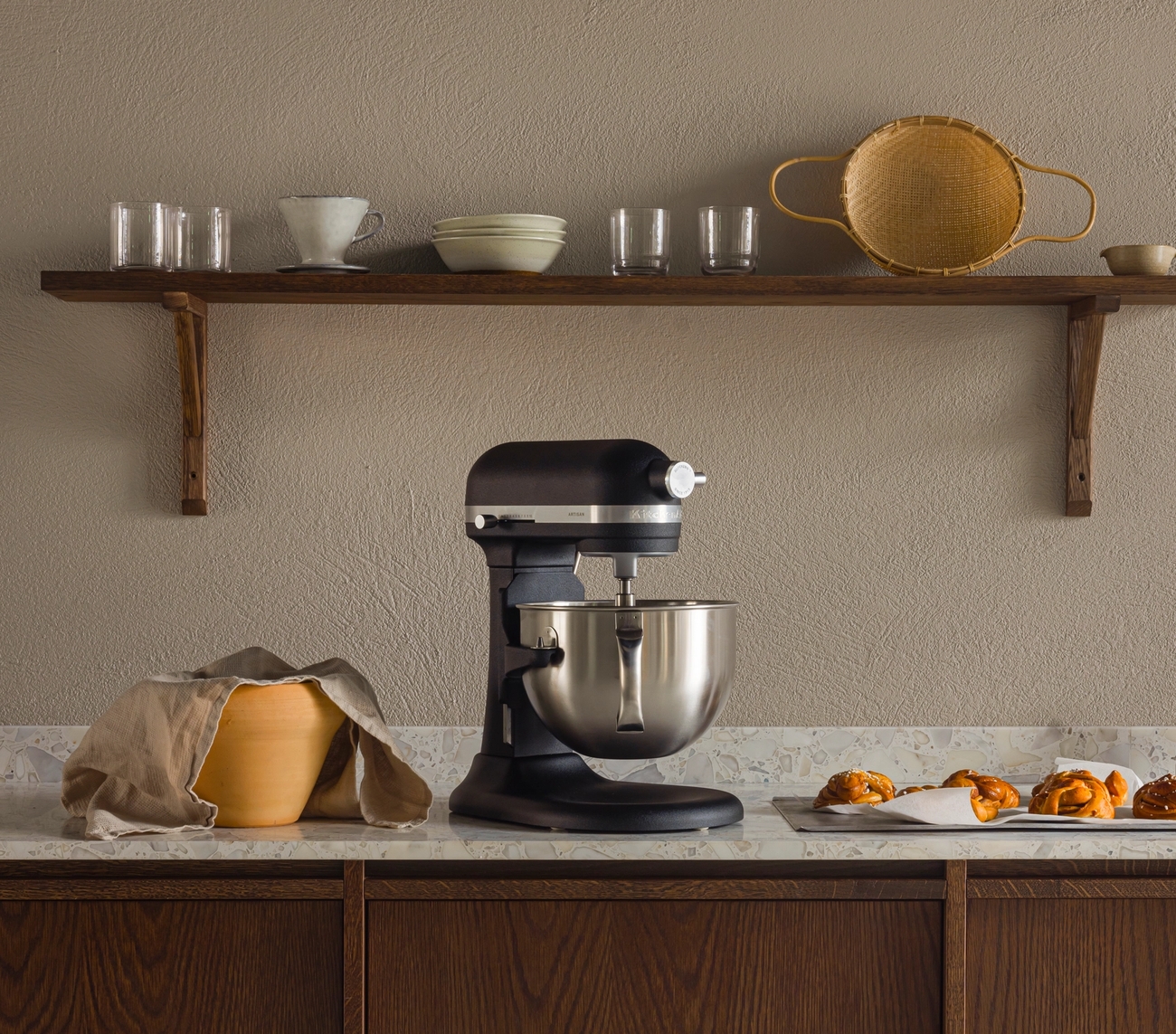 En kjøkkenbenk med en svart kjøkkenmaskin, en tildekket deigbolle og bakverk på et brett, under en trehylle med kjøkkenutstyr.
