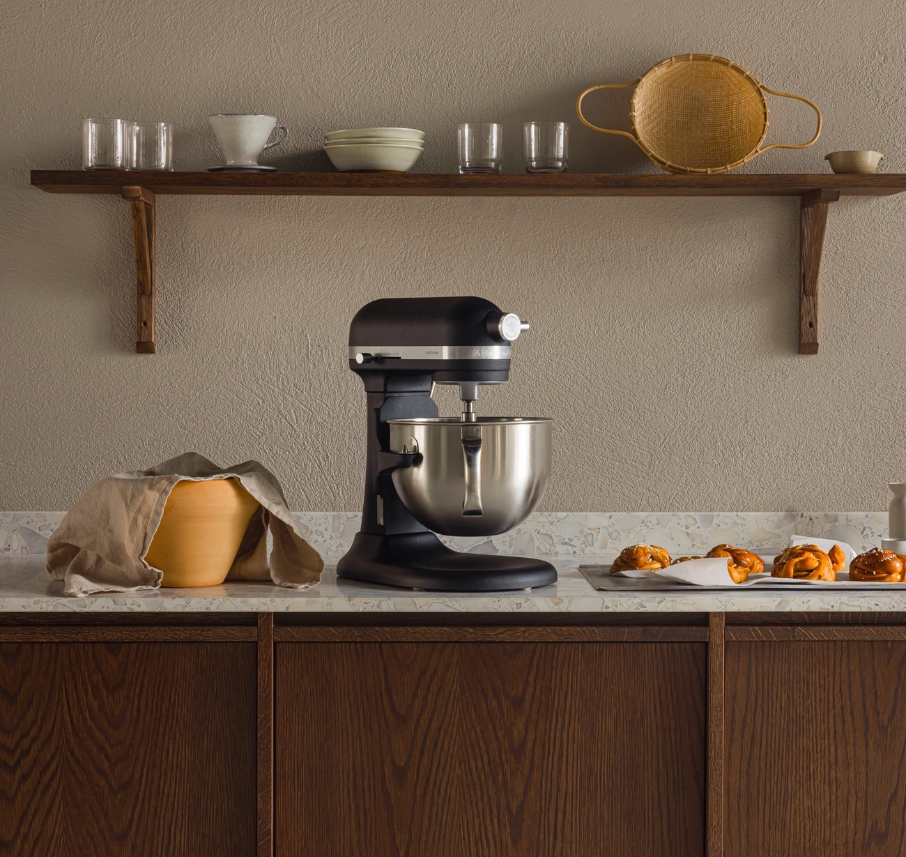 En köksbänk med en svart köksmaskin, en täckt degbunke och bakverk på en bricka, under en trähylla med köksredskap.