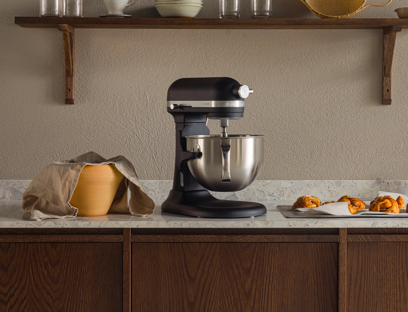 Keittiön tiskillä musta yleiskone, kansillinen taikinakulho ja leivonnaisia tarjottimella puisen keittiövälineitä esittelevän hyllyn alla.