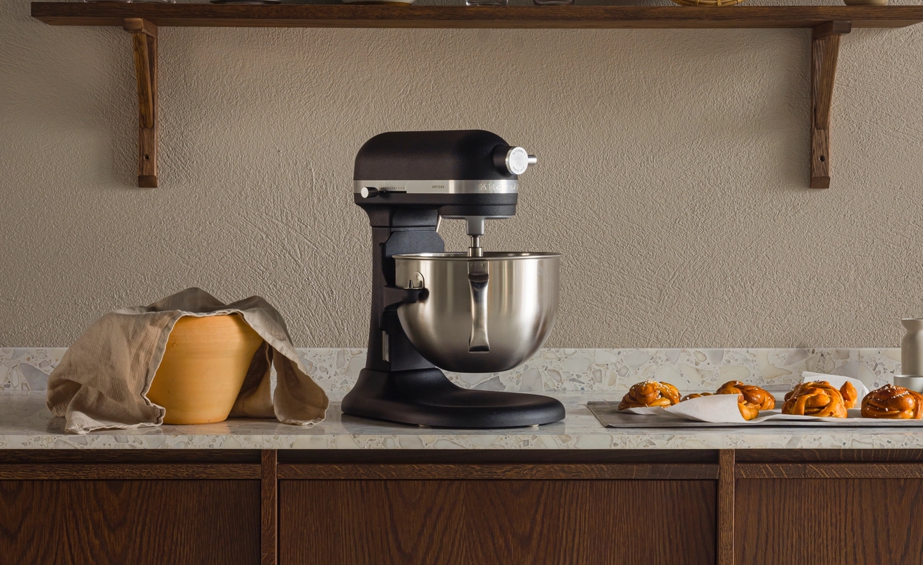 En kjøkkenbenk med en svart kjøkkenmaskin, en tildekket deigbolle og bakverk på et brett, under en trehylle med kjøkkenutstyr.