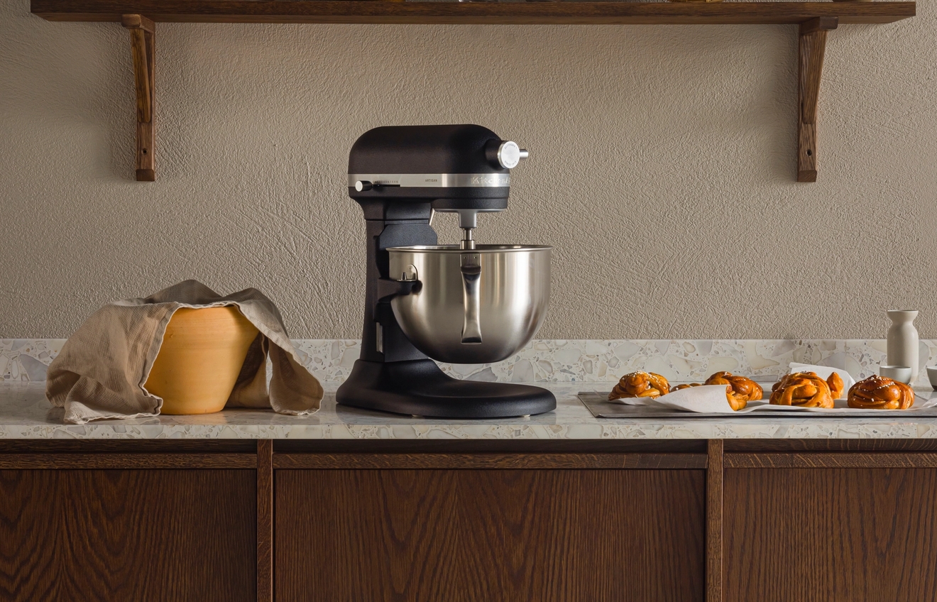 Keittiön tiskillä musta yleiskone, kansillinen taikinakulho ja leivonnaisia tarjottimella puisen keittiövälineitä esittelevän hyllyn alla.