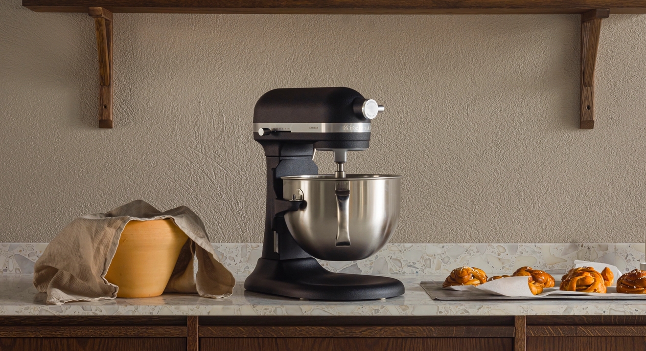 En köksbänk med en svart köksmaskin, en täckt degbunke och bakverk på en bricka, under en trähylla med köksredskap.