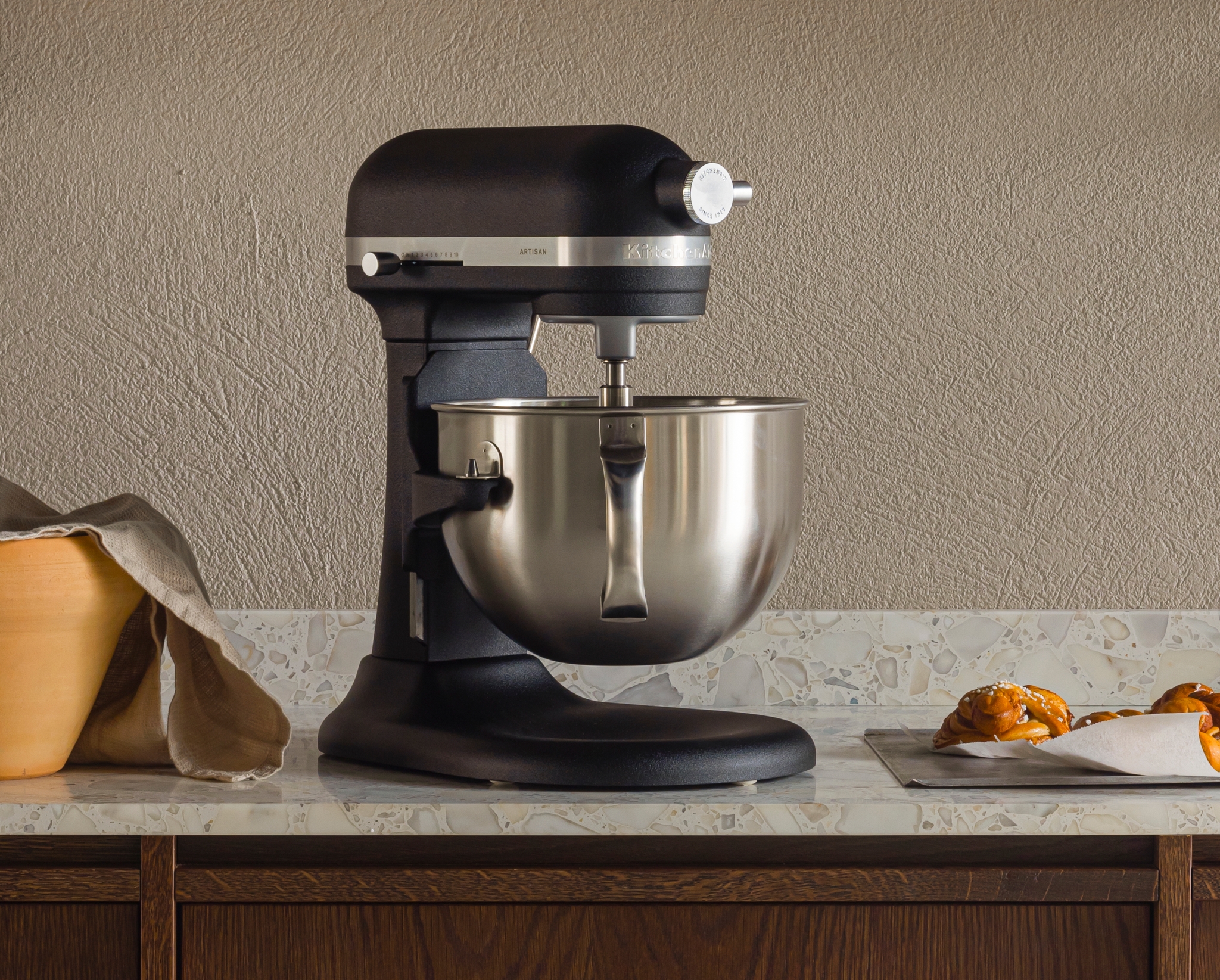 Moderni keittiö, jossa on tummat puukaapit, yleiskone ja leivonnaisia vaalealla työtasolla sekä astiahylly.