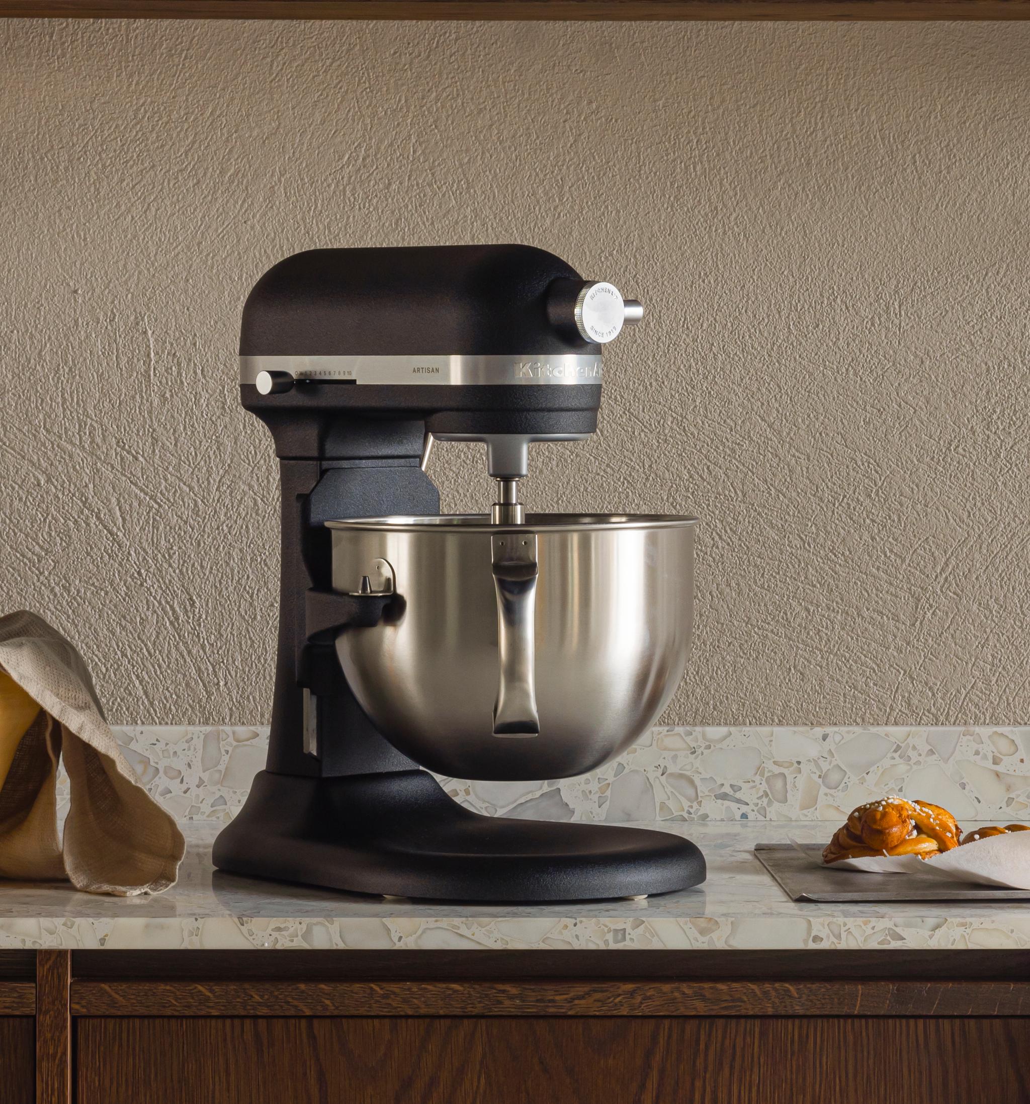 Moderni keittiö, jossa on tummat puukaapit, yleiskone ja leivonnaisia vaalealla työtasolla sekä astiahylly.