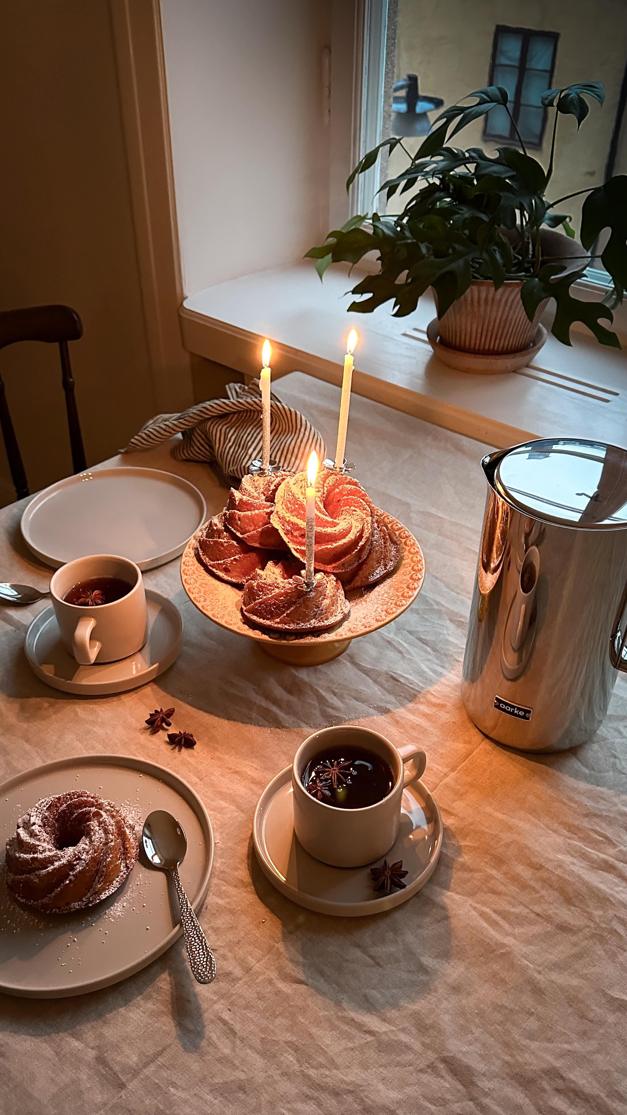A cozy table setting with lit candles on bundt cakes, mugs of hot drinks garnished with star anise, and a silver pitcher.