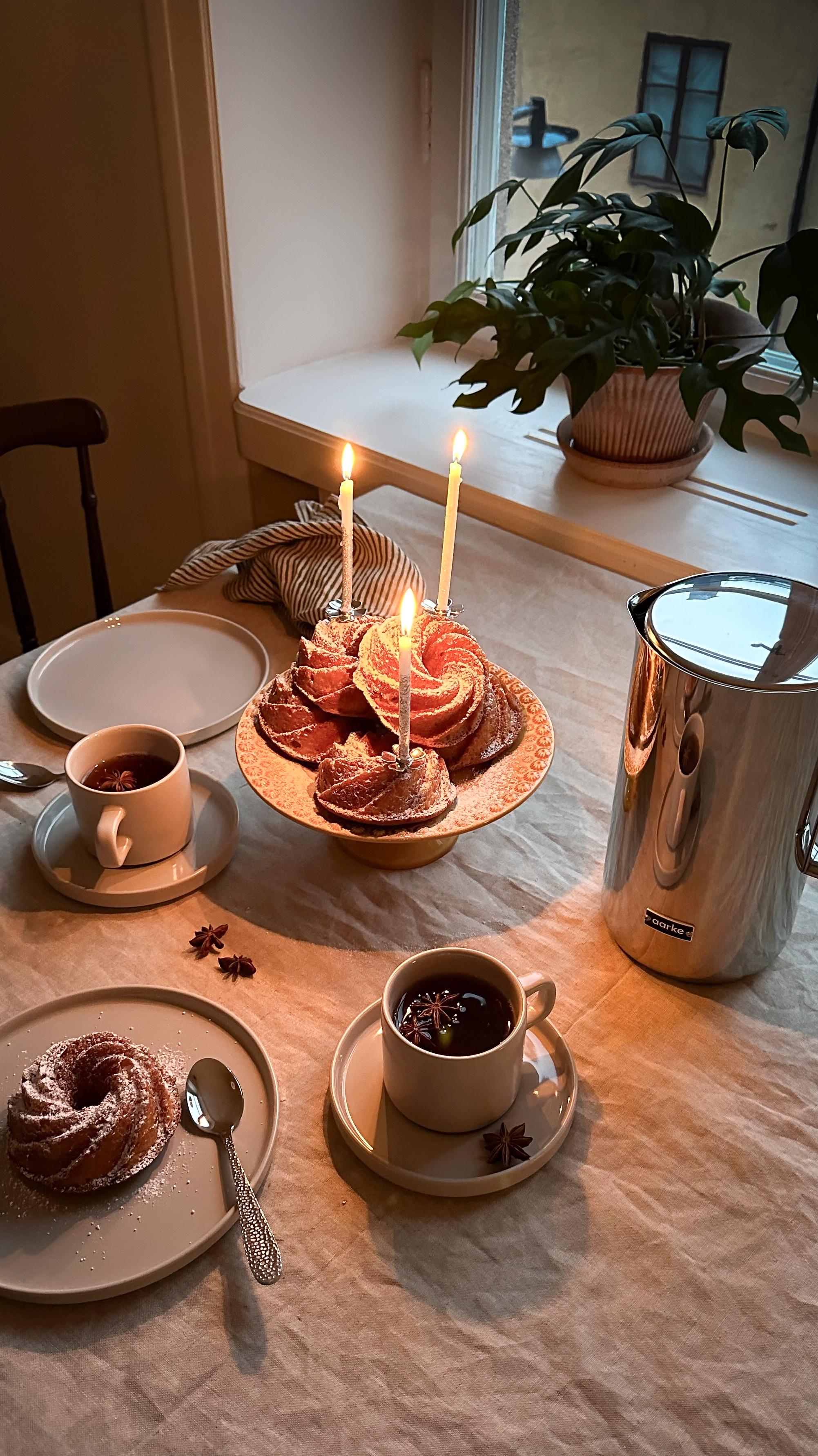 A cozy table setting with lit candles on bundt cakes, mugs of hot drinks garnished with star anise, and a silver pitcher.