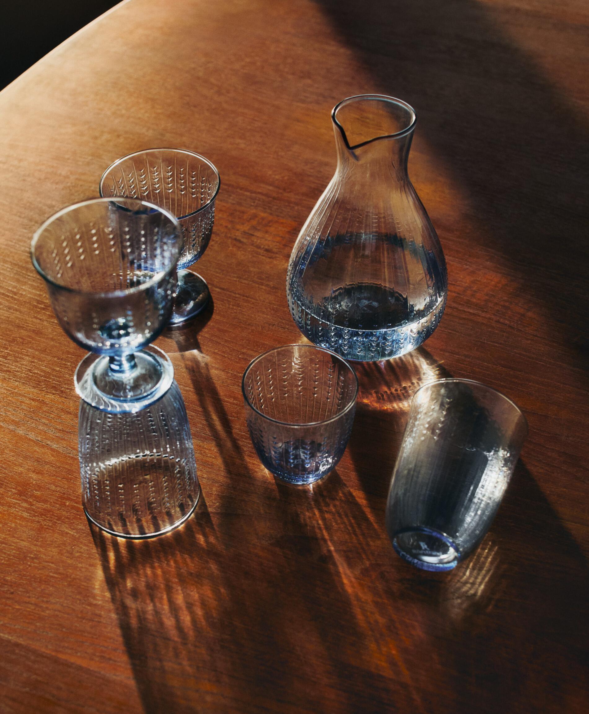 Blå teksturert glasskaraffel og glass på et trebord med sterke skygger.