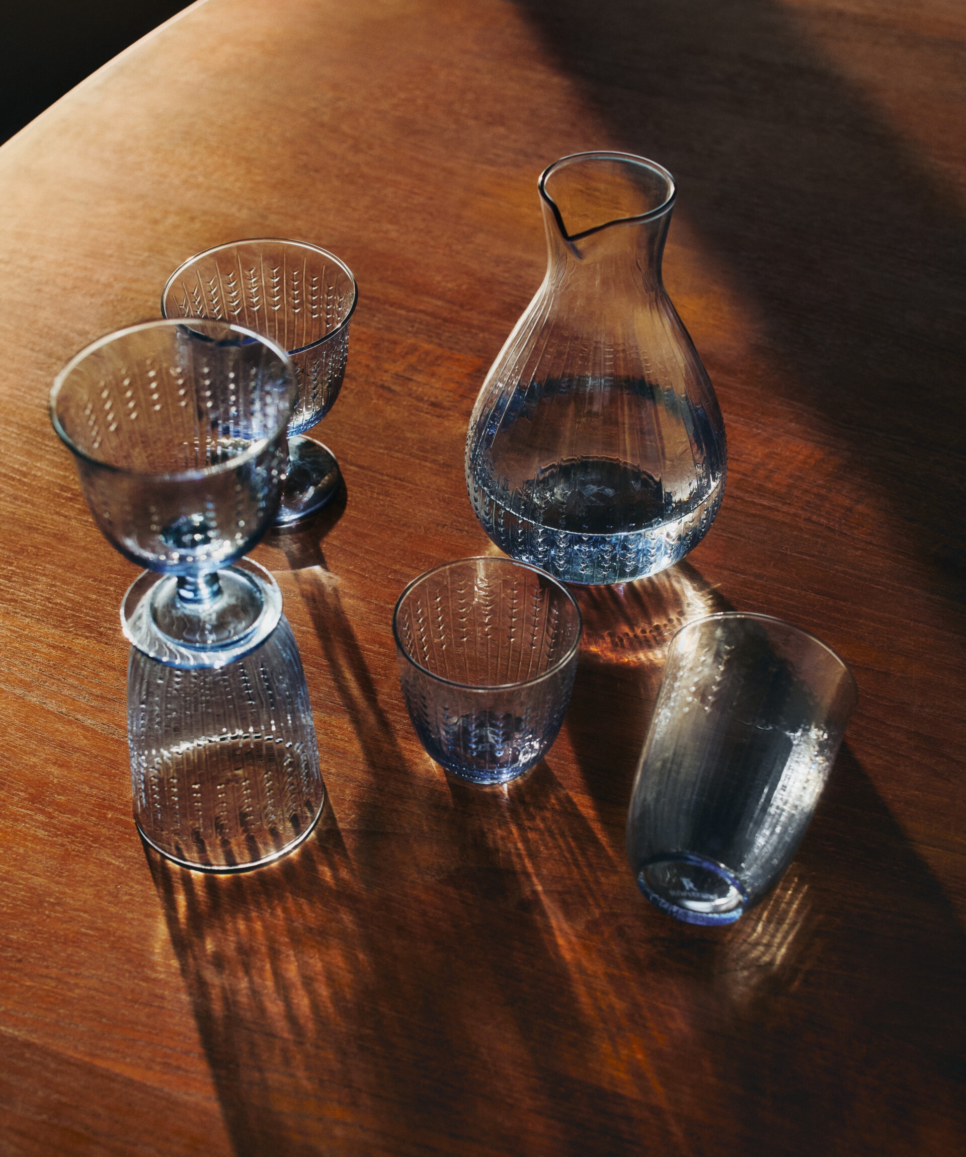 Blå teksturert glasskaraffel og glass på et trebord med sterke skygger.