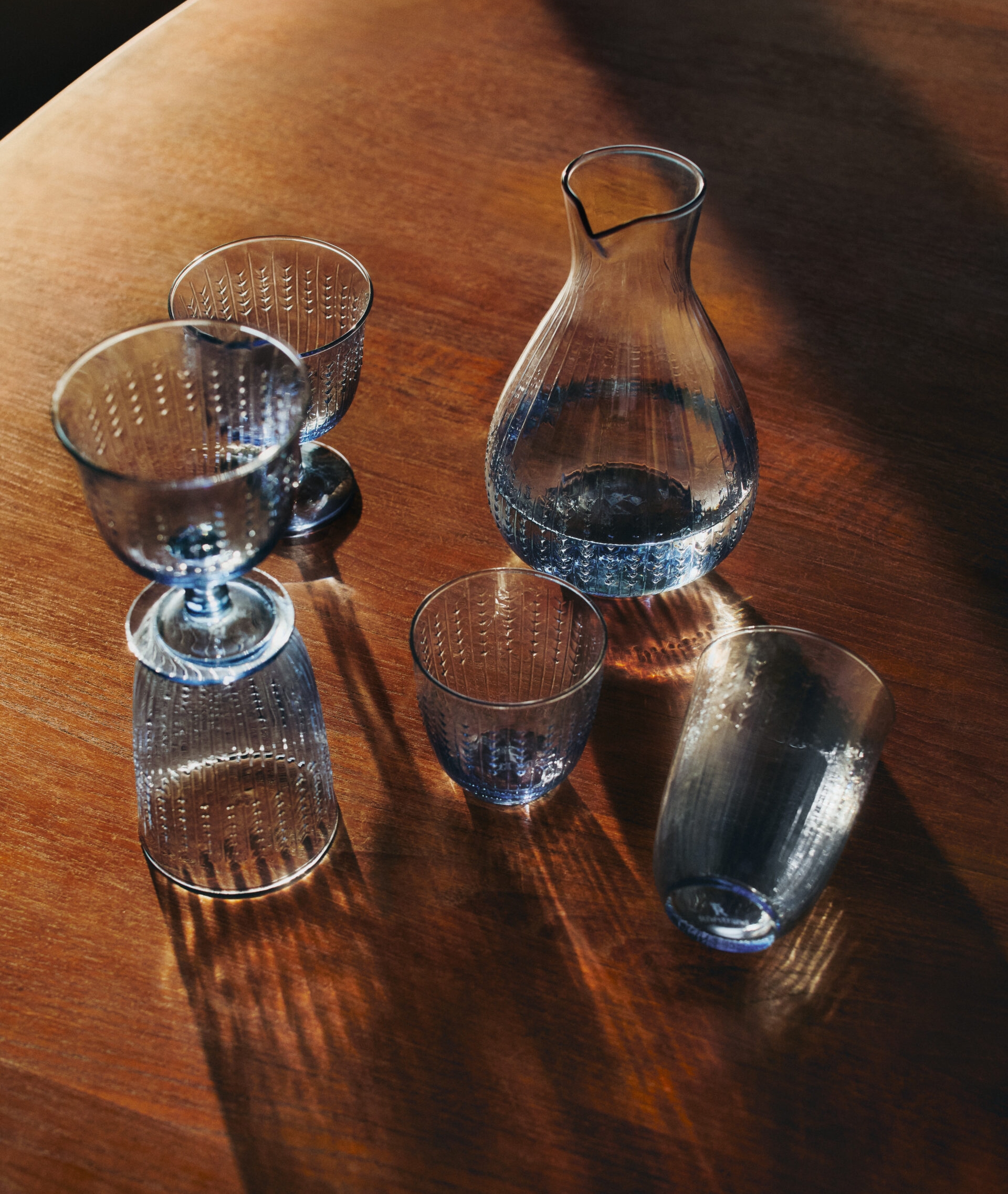 Blå texturerad glaskaraff och glas på ett träbord med starka skuggor.