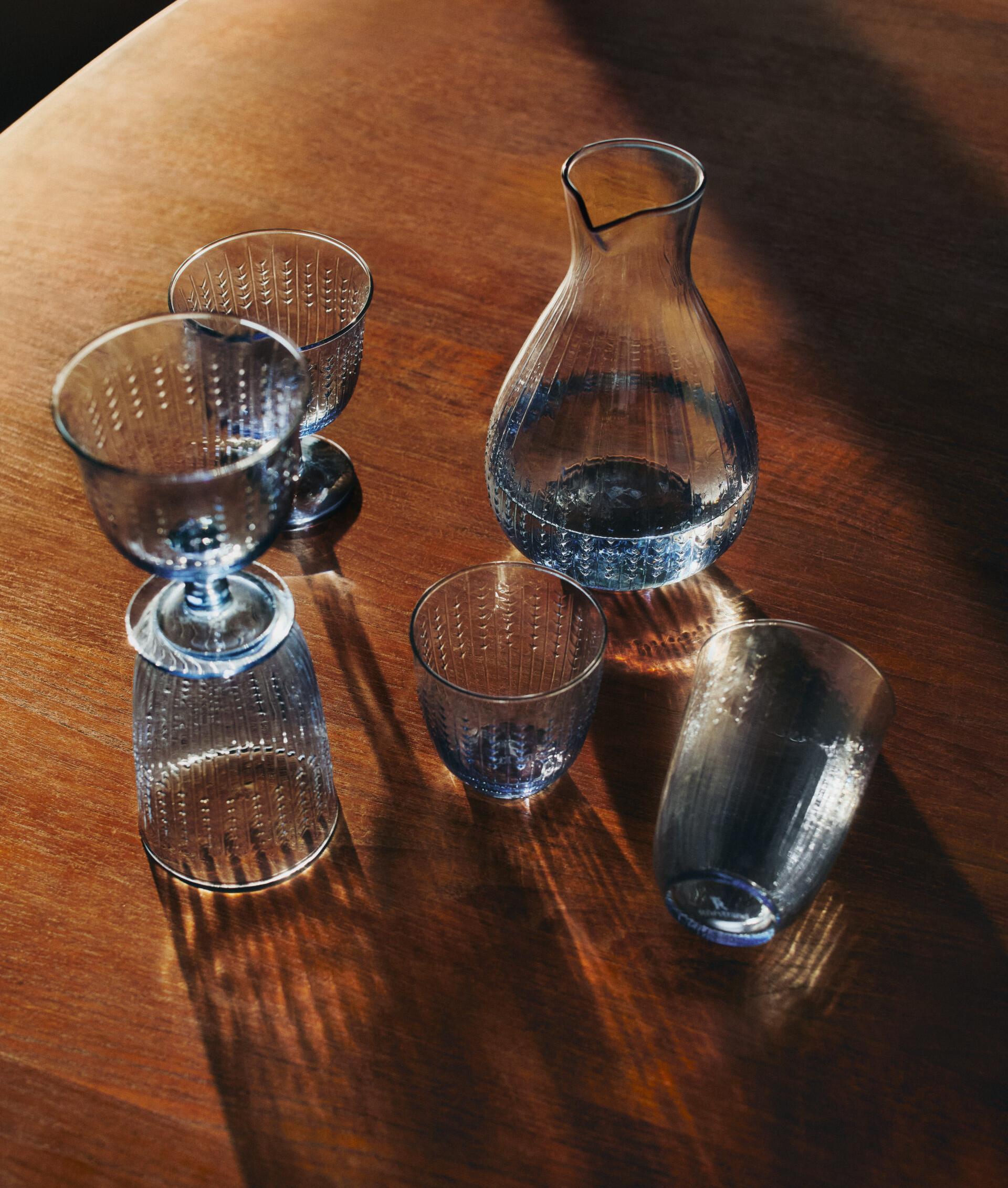 Blå texturerad glaskaraff och glas på ett träbord med starka skuggor.