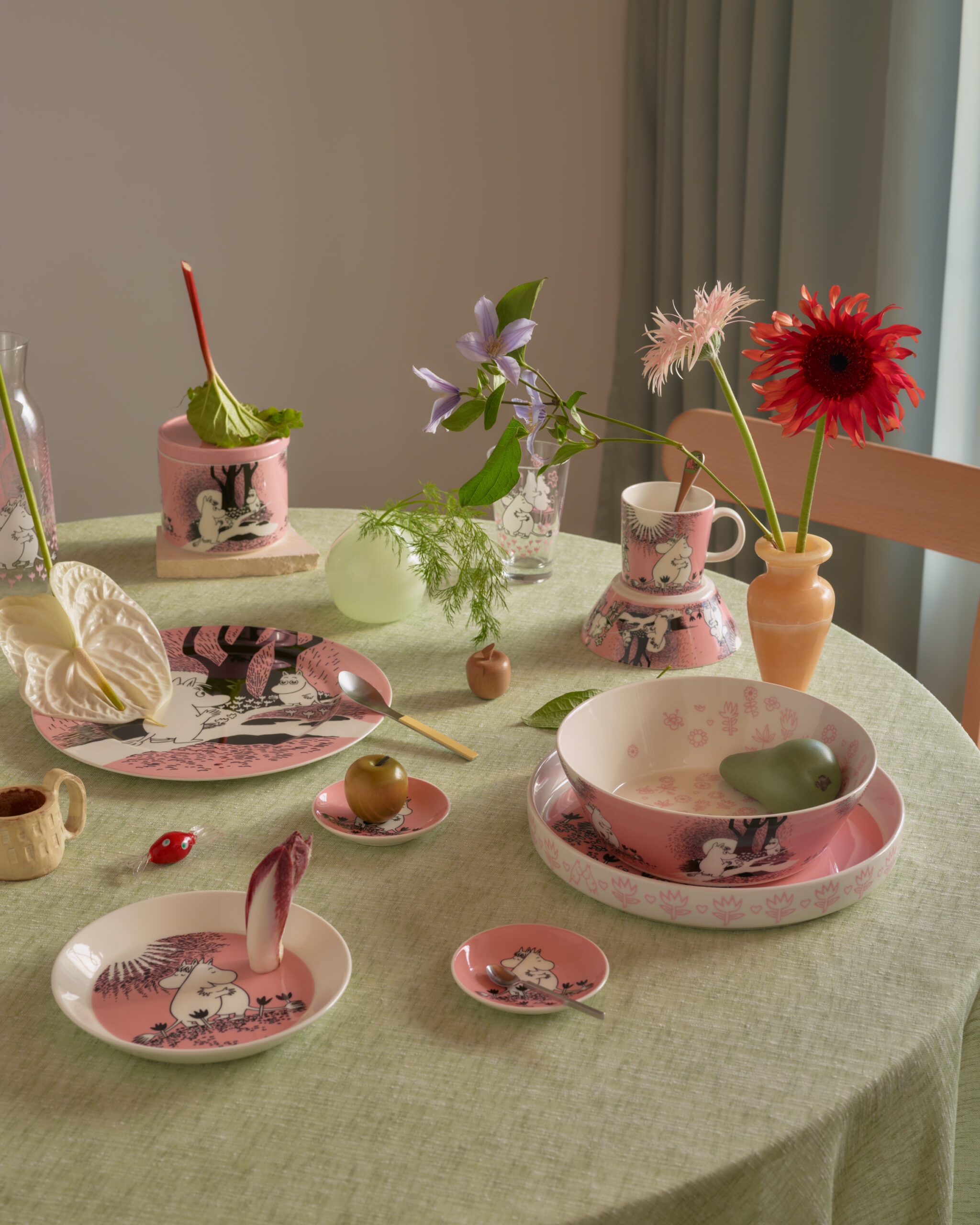 Et dekket bord med rosa og hvitt Mummi-servise, dekorative blomster og små frukter på en grønn duk.