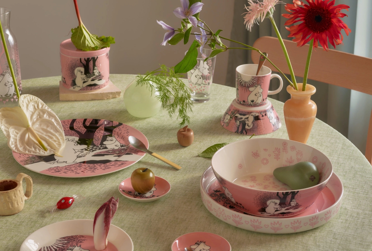 Rosa og hvitt mummitema-servise, inkludert tallerkener og skåler, plassert på en lysegrønn duk med en blomst, eple og pære