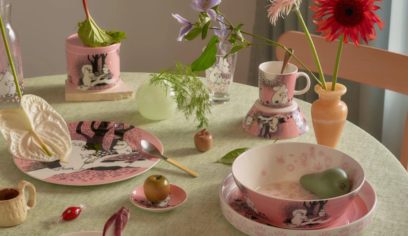 Rosa og hvitt mummitema-servise, inkludert tallerkener og skåler, plassert på en lysegrønn duk med en blomst, eple og pære