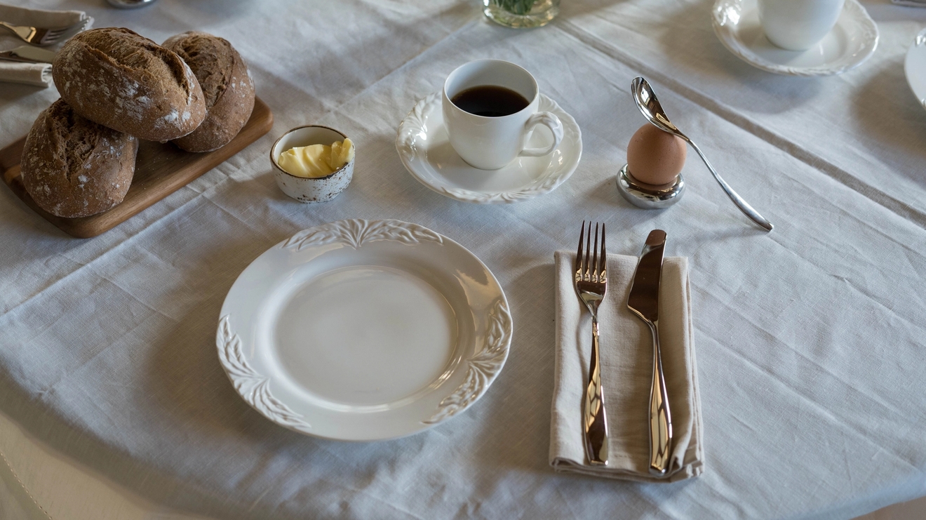 Pöytä katettu aamiaiseksi lautasilla, kupeilla, aterimilla ja leivällä.