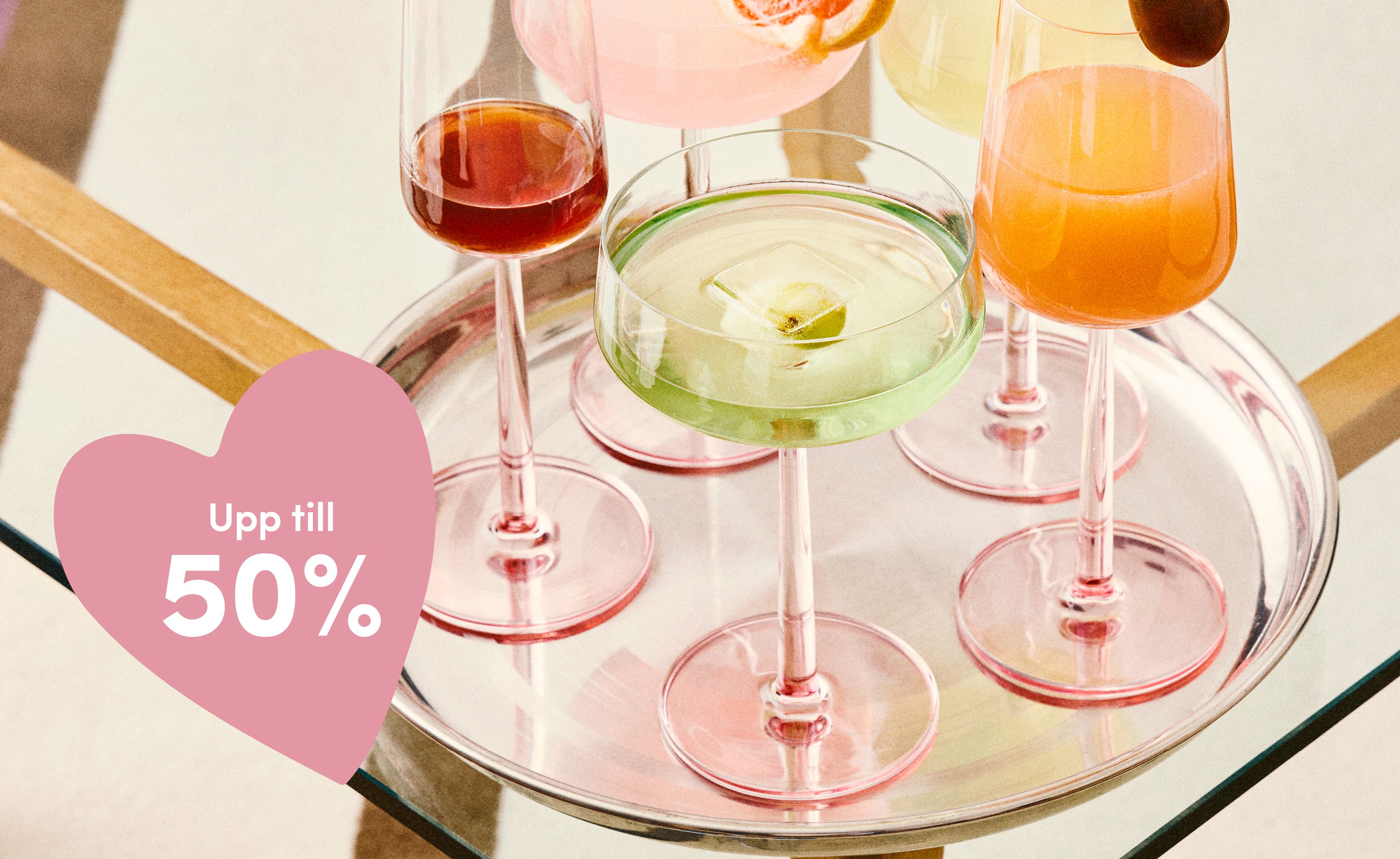 Blandade cocktails i glas med rosa fot på en bricka med en hjärtformad text "Upp till 50 %" rabatt.