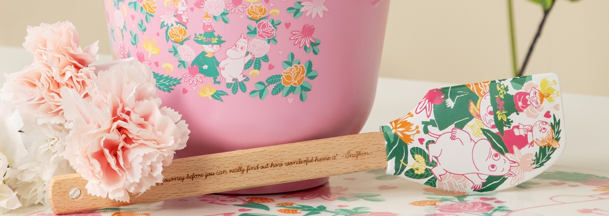 Vaaleanpunainen kulho ja nuolija muumikuviolla ovat pöydällä kukkien vieressä.