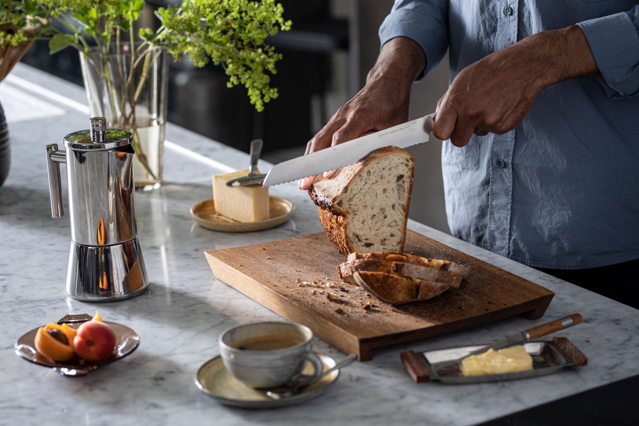 Kädet viipaloivat leipää puulaudalla, kahvinkeitin, kahvikuppi, voi, juusto ja hedelmät marmorisella tiskillä.