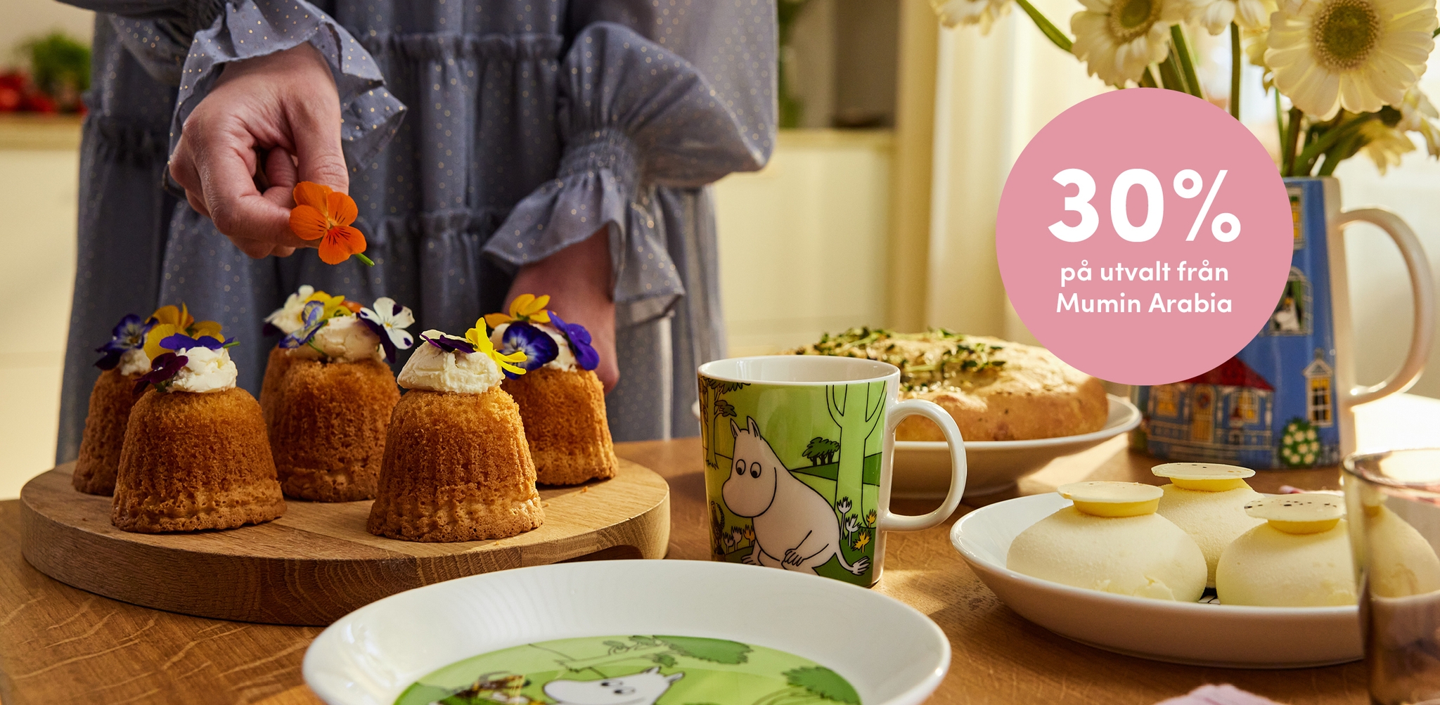 En person dekorerar små kakor med ätbara blommor på ett bord dukat med Mumin-rätter och andra desserter, med ett rosa överlägg som annonserar "30 % rabatt på utvalda Mumin Arabia-produkter".