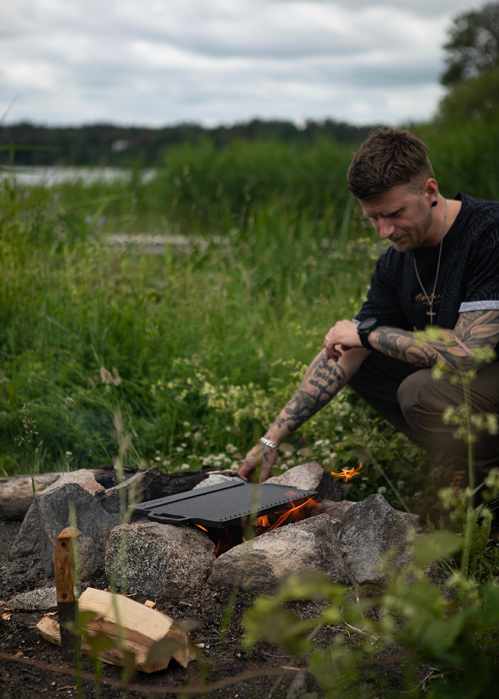 Man lagar mat över öppen eld på en grillplatta vid en stenring ute i naturen.