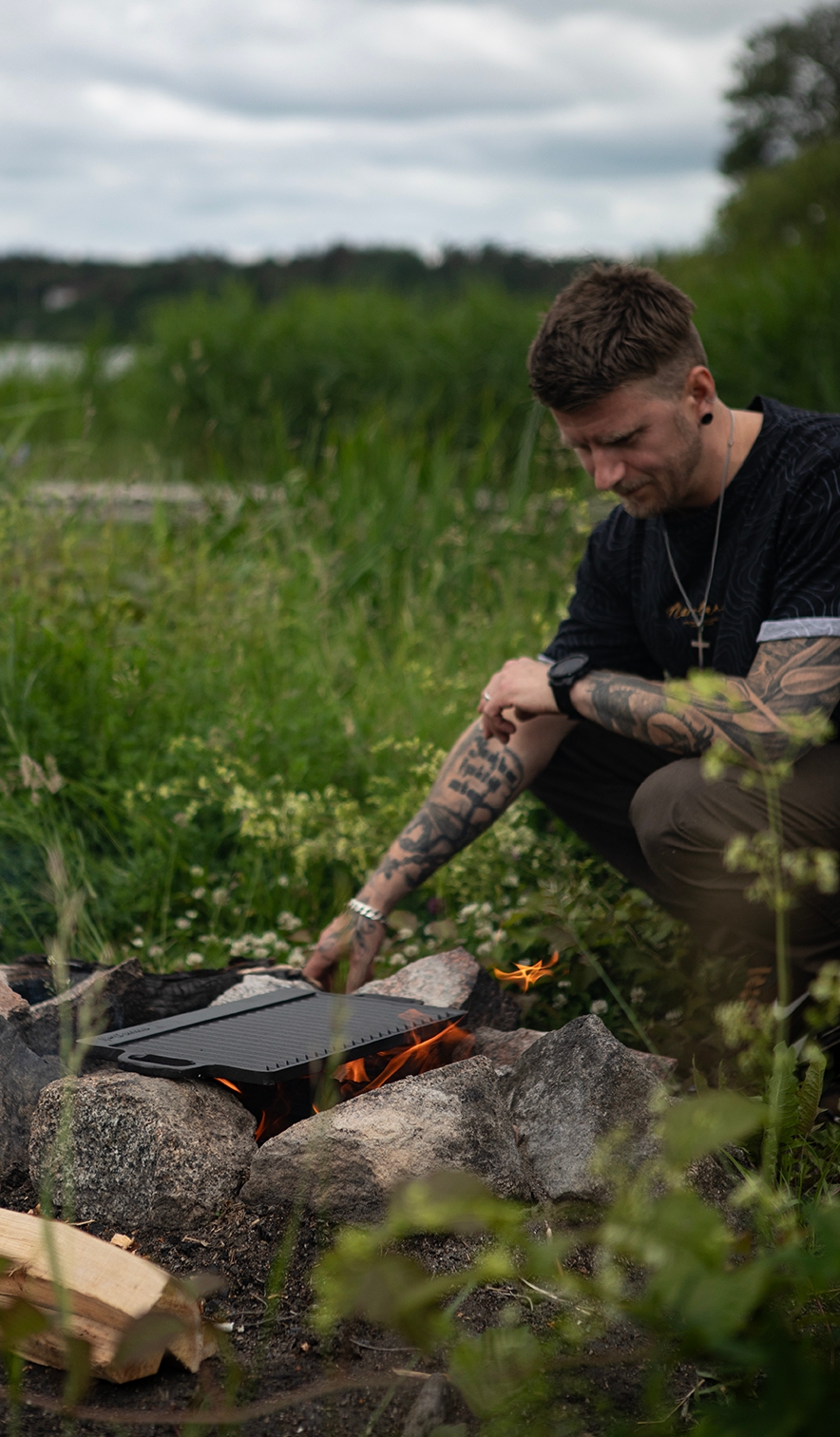 Man grillar över öppen eld vid en stenring ute i naturen intill sjö och grönska.
