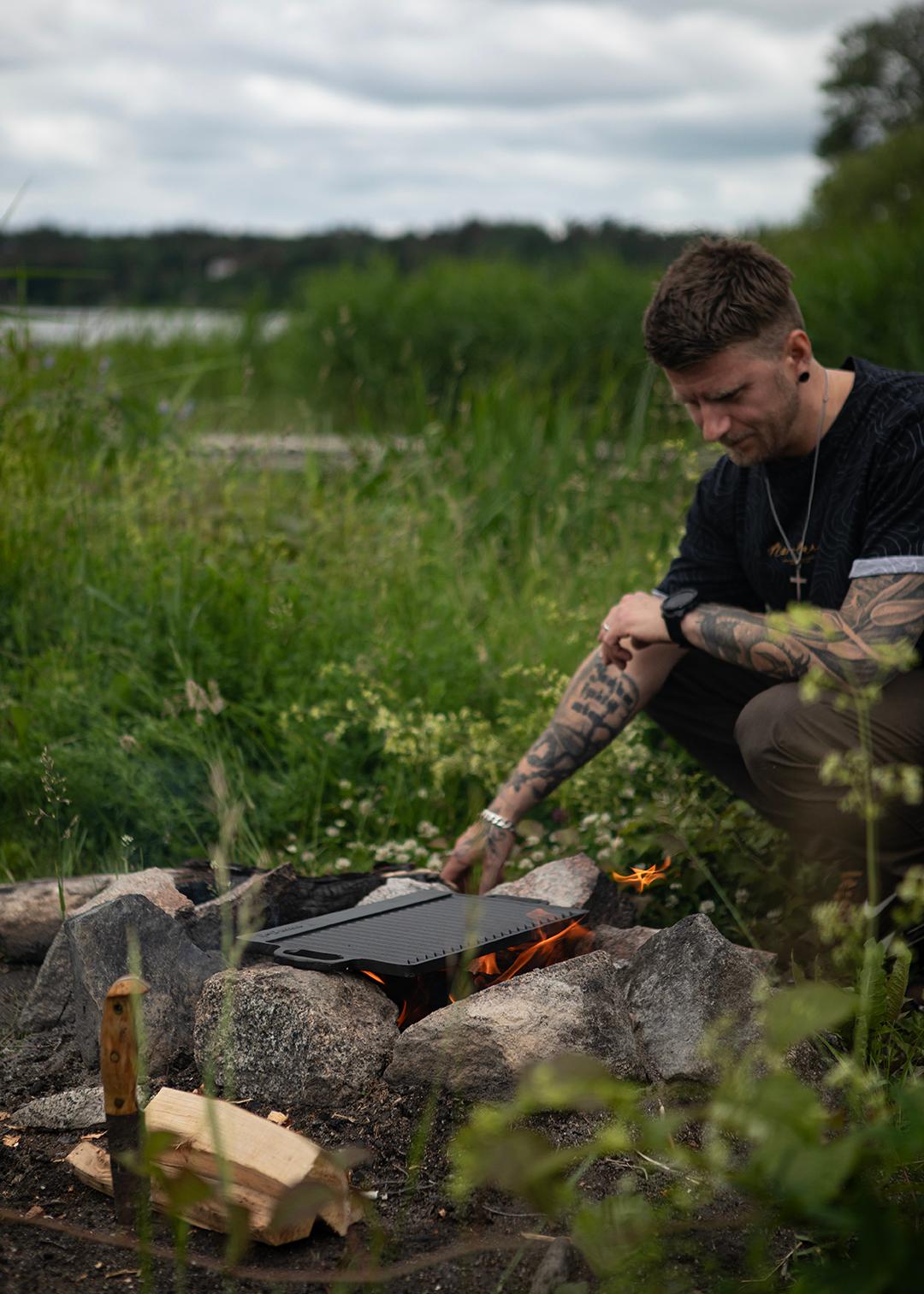 Man lagar mat över öppen eld på en grillplatta vid en stenring ute i naturen.