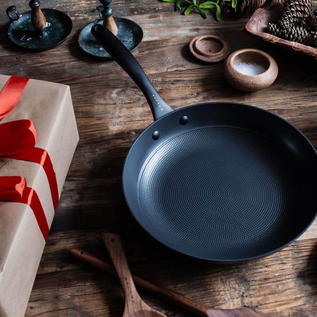 En mörkgrå stekpanna med strukturerad botten står på ett rustikt träbord bredvid en brun papperspresent knuten med ett rött band.