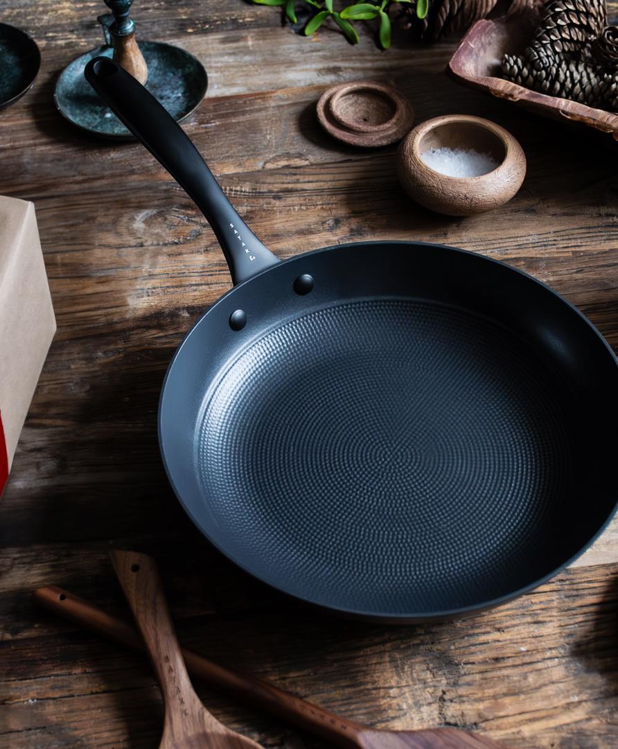 En mörkgrå stekpanna med strukturerad botten står på ett rustikt träbord bredvid en brun papperspresent knuten med ett rött band.