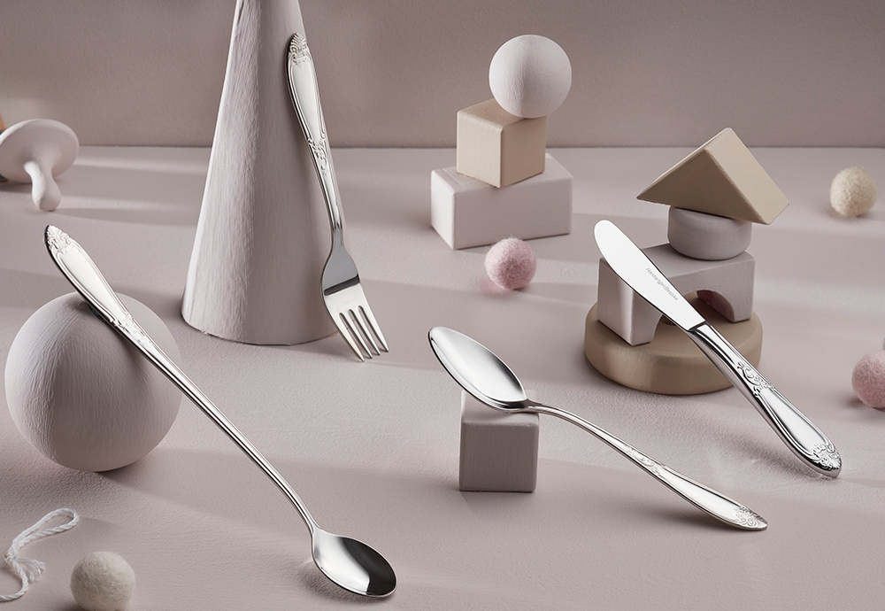 Elegant silver child's cutlery (spoon, fork, knife) on a pastel pink surface with decorative blocks and balls.