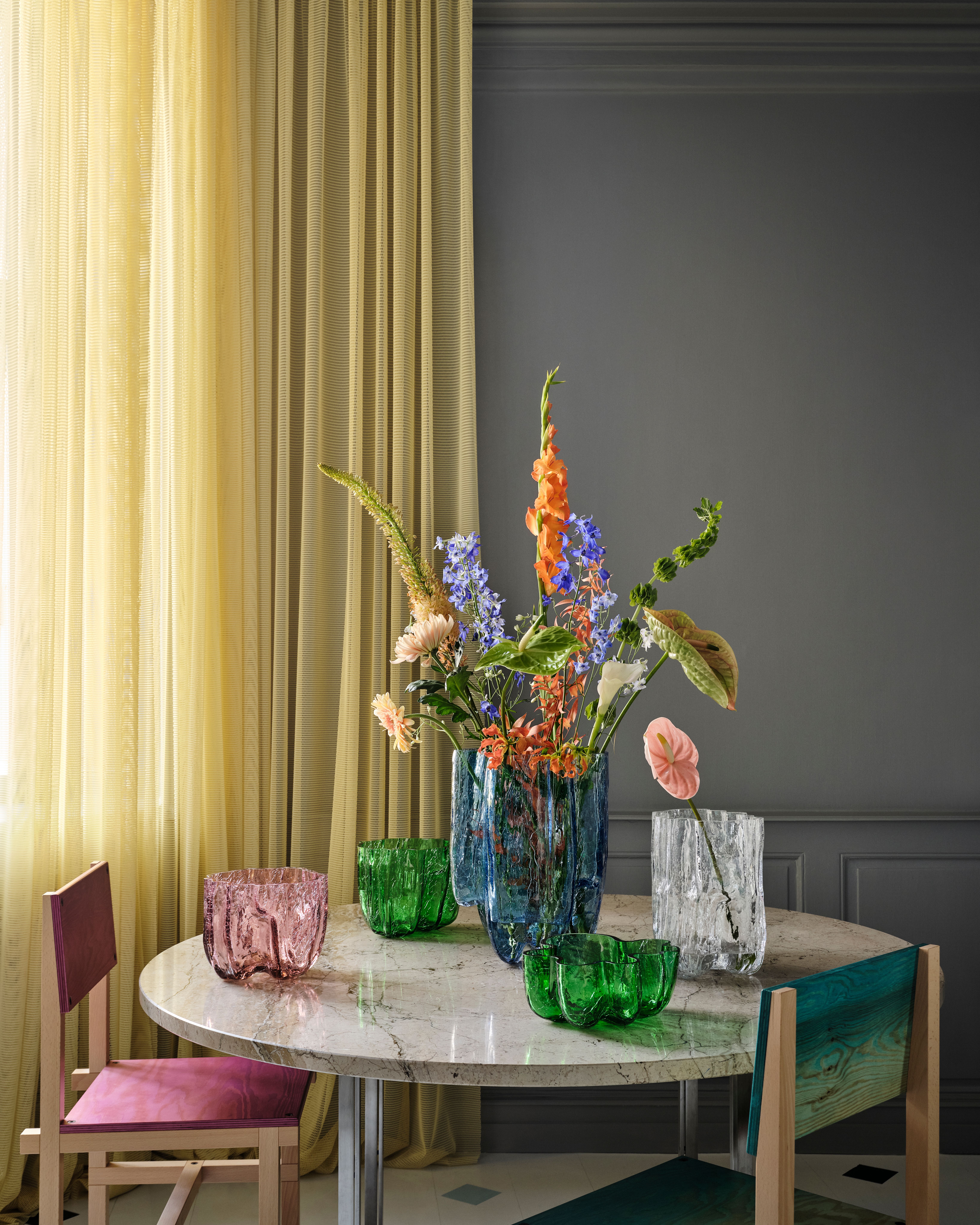 A marble table displays colorful flowers in a blue textured vase, with smaller green, pink, and clear textured glass vases, and two colorful chairs.