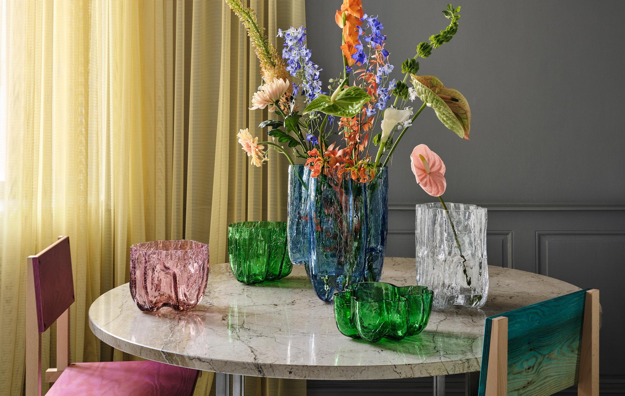 En samling texturerade glasvaser och skålar i blått, grönt, rosa och genomskinligt, några med blommor, på ett bord.