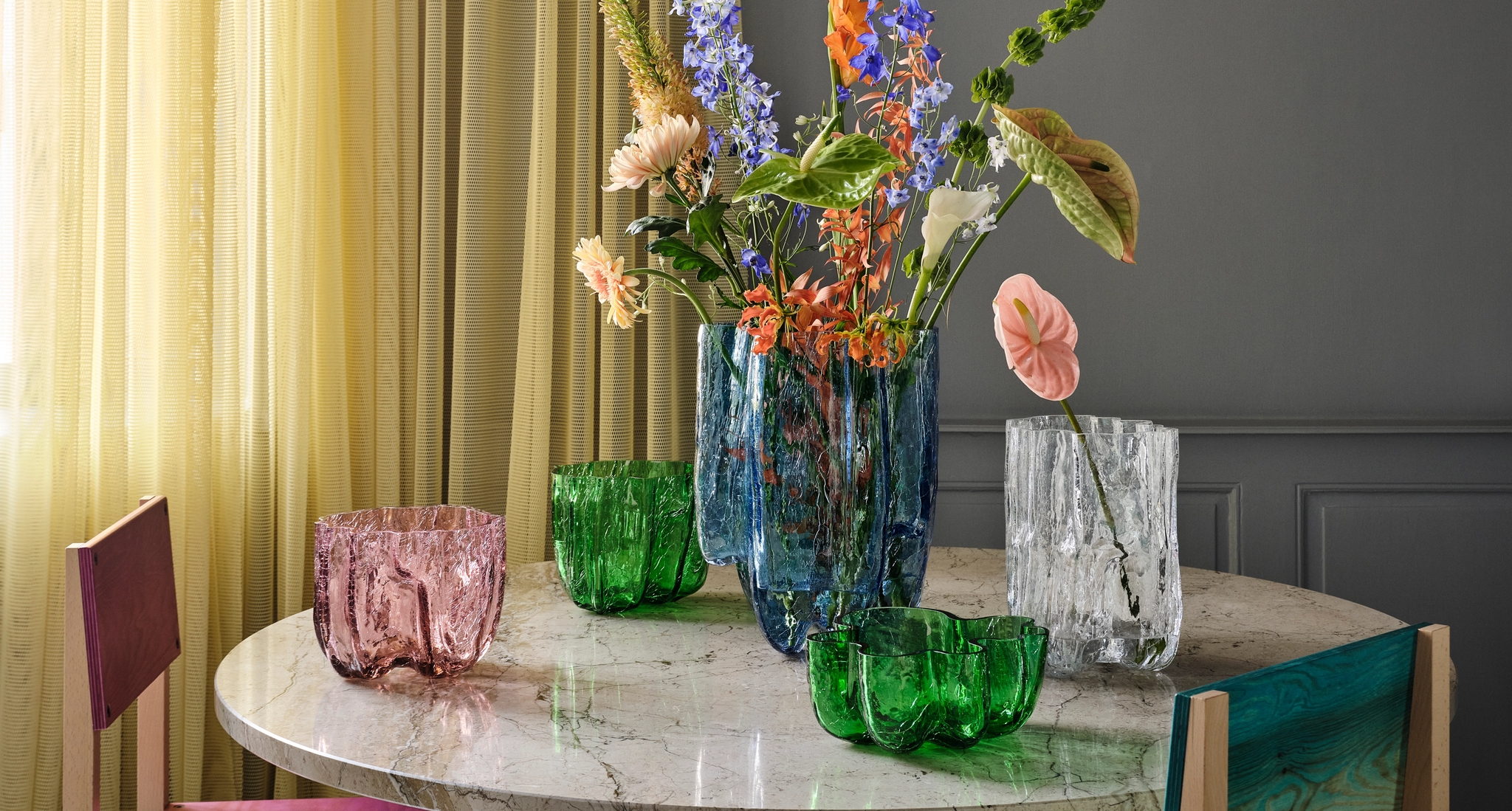 En samling texturerade glasvaser och skålar i blått, grönt, rosa och genomskinligt, några med blommor, på ett bord.