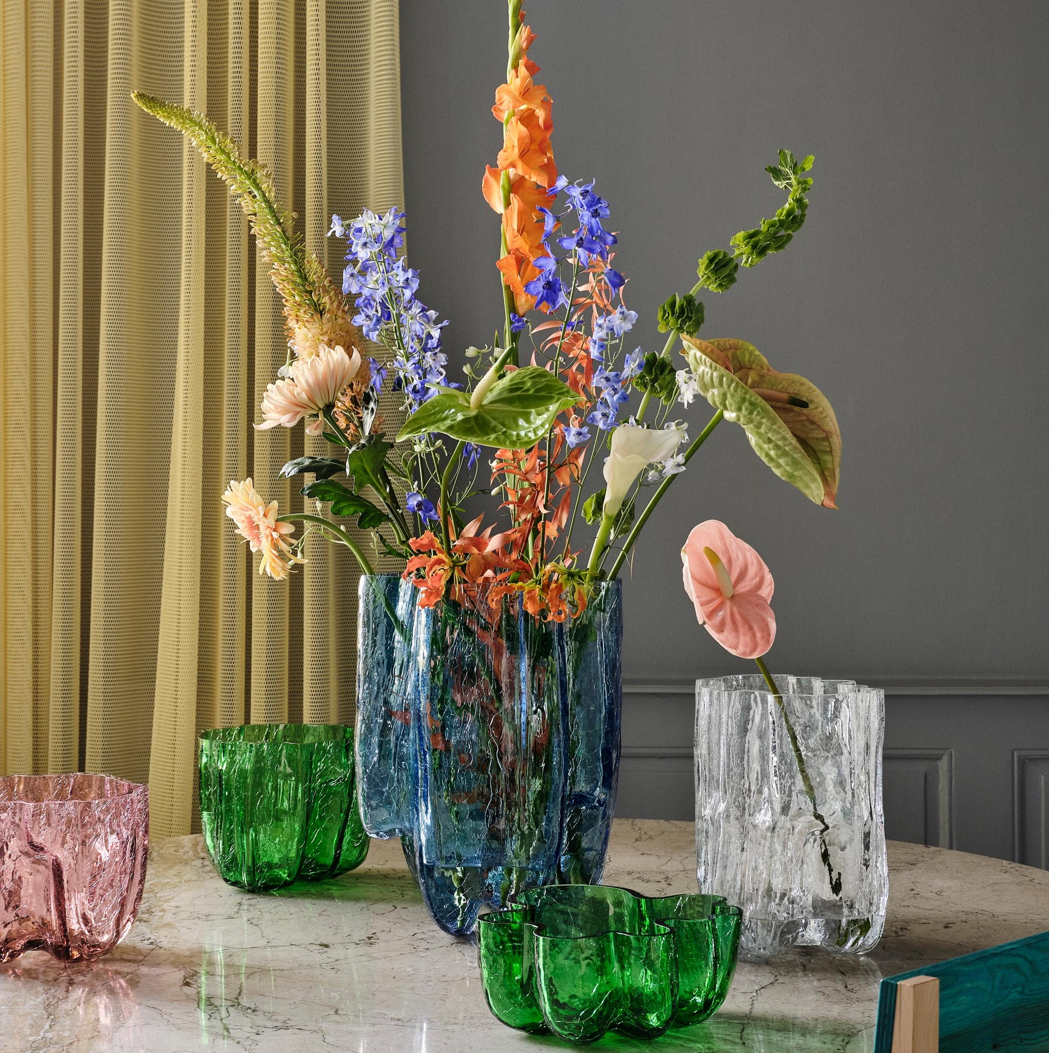 A marble table displays colorful flowers in a blue textured vase, with smaller green, pink, and clear textured glass vases, and two colorful chairs.