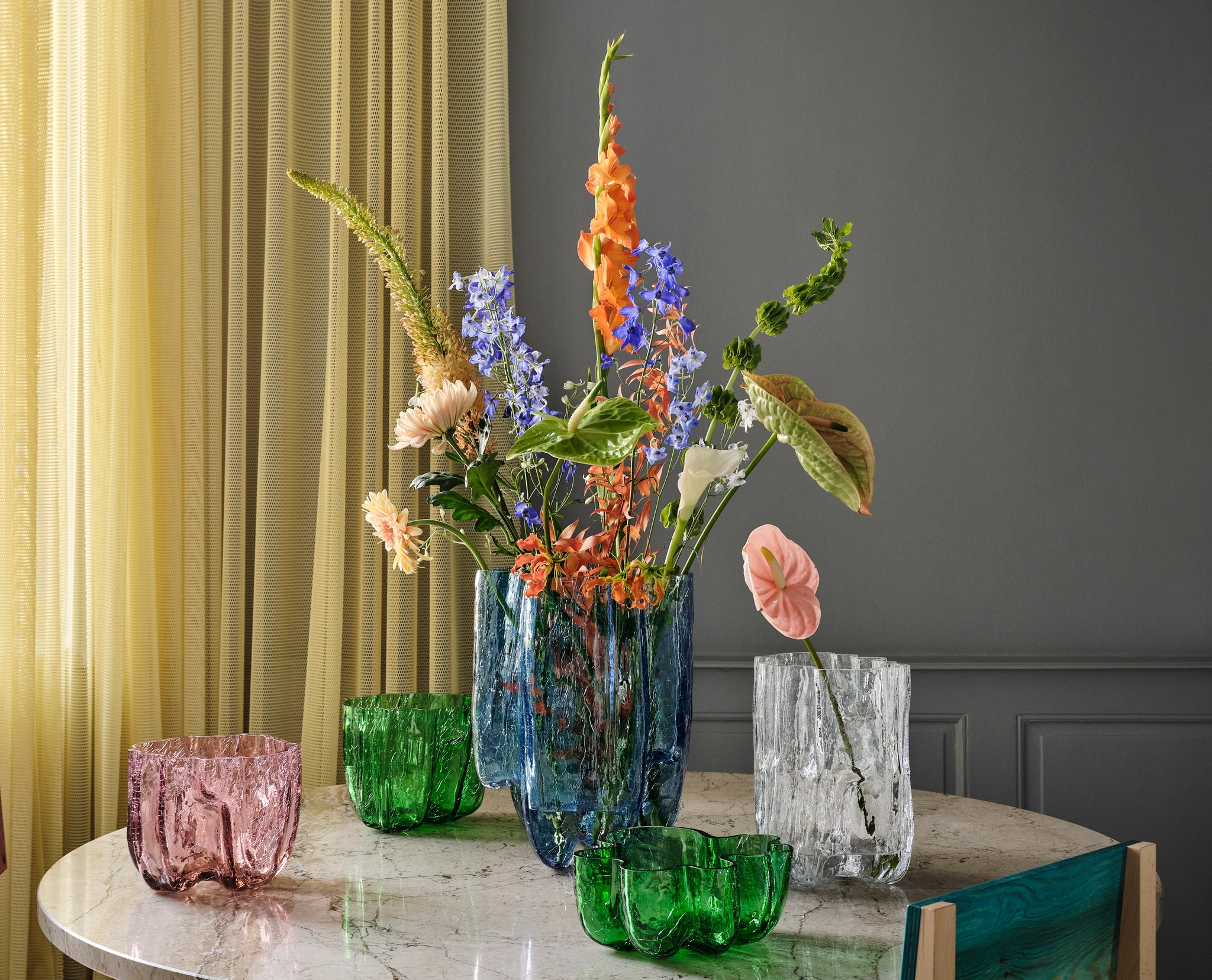 A round marble table features a large blue vase with colorful flowers, surrounded by smaller green, pink, and clear glass vases, with a pink chair and yellow curtains in the background.