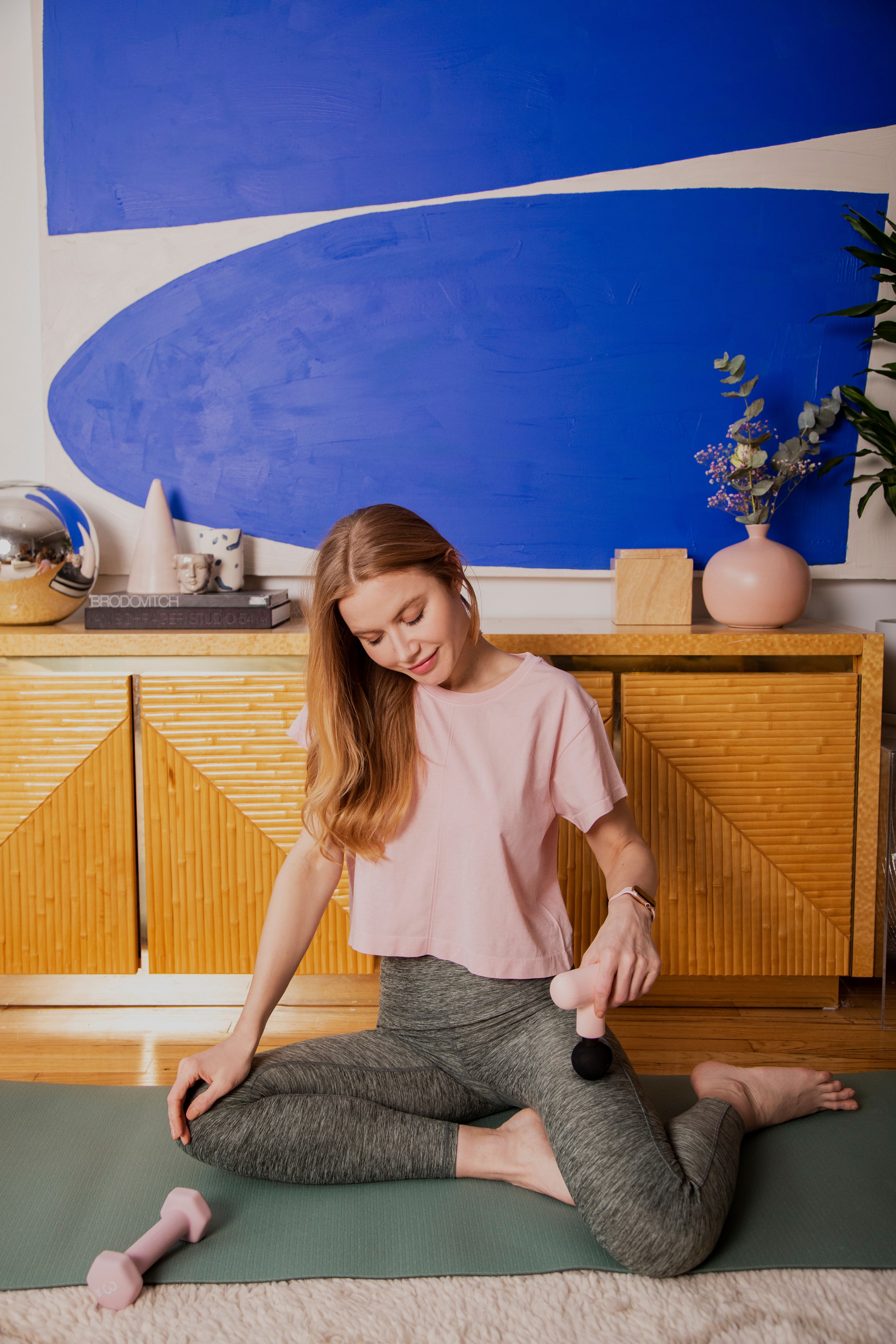 Kvinna i träningskläder sitter på yogamatta och masserar benet med en massagepistol i ett modernt vardagsrum.