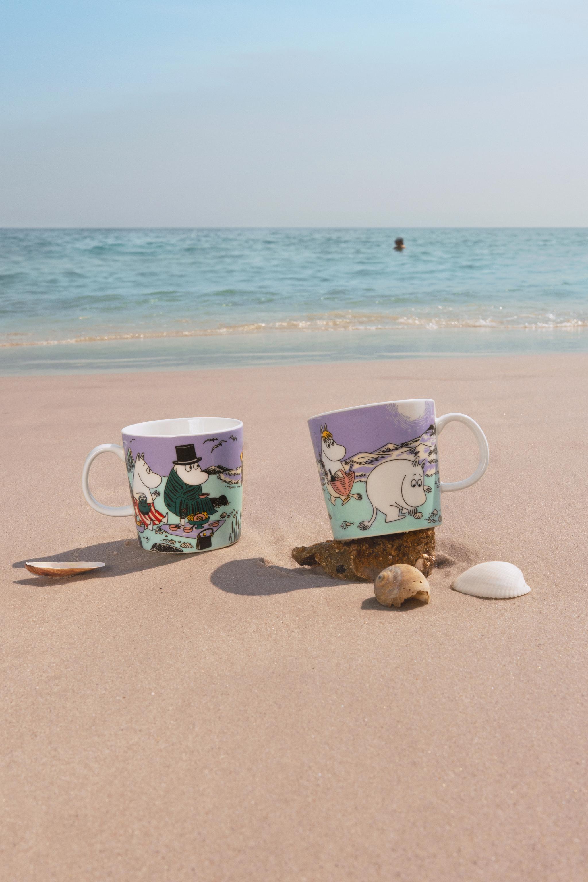 two mugs are sitting on a sandy beach near the ocean