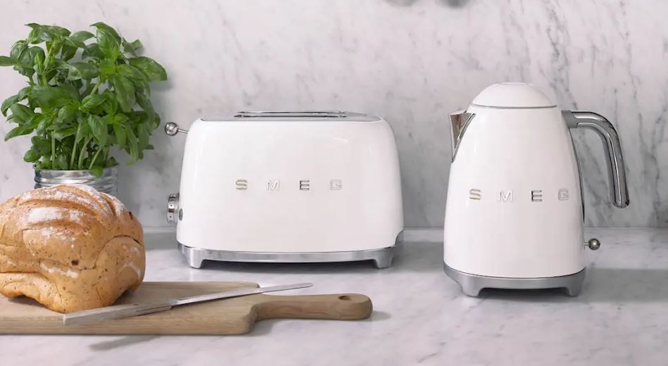 Hvit Smeg-brødrister og vannkoker på en kjøkkenbenk i marmor med fersk basilikum, brød og kjøkkenutstyr i kobber hengende over.