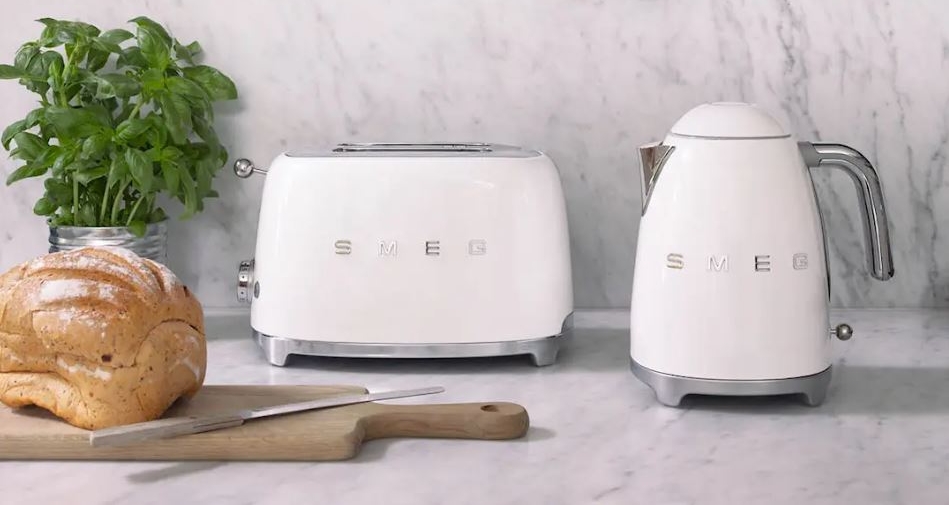Stilren kjøkkenbenk med hvit SMEG-brødrister og vannkoker, ferskt brød og urter foran marmorvegg.