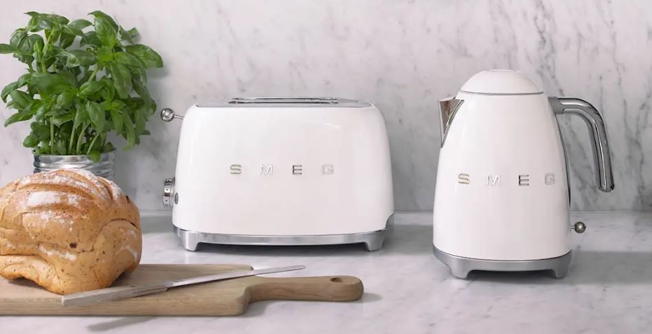 Vit Smeg-brödrost och vattenkokare på en marmorköksbänk med färsk basilika, bröd och köksredskap i koppar som hänger ovanför.