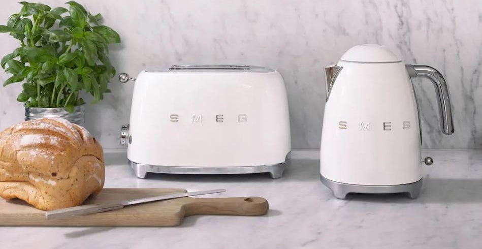 Valkoinen Smeg-leivänpaahdin ja vedenkeitin marmoritasolla, jossa on tuoretta basilikaa, leipää ja yläpuolella roikkuvia kuparisia keittiövälineitä.