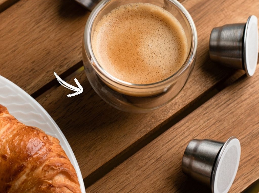 A cup of espresso, reusable coffee capsules, and a croissant on a wooden table. Text: MAXIMUM FLAVOR EXTRACTION.