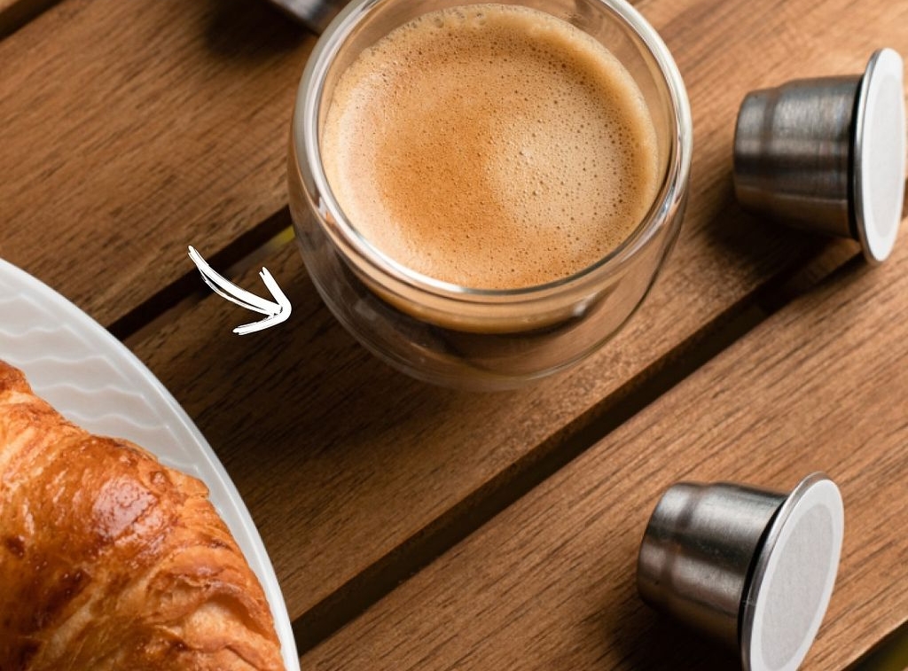 Espresso in a glass cup, reusable metal coffee pods, and a croissant on a wooden table. Text: "MAXIMUM FLAVOR EXTRACTION."